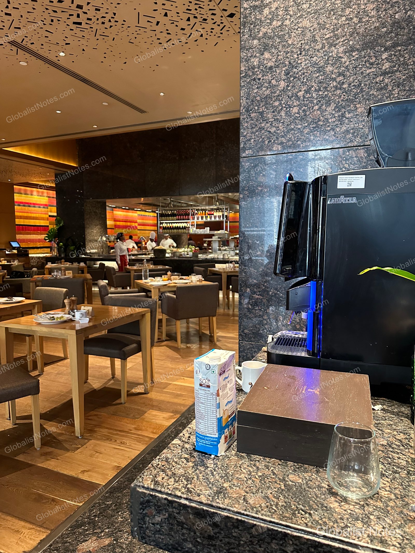 The breakfast restaurant at Grand Hyatt Mumbai, featuring a Lavazza coffee machine and soy milk in the foreground, with an open kitchen and dining area in the background.