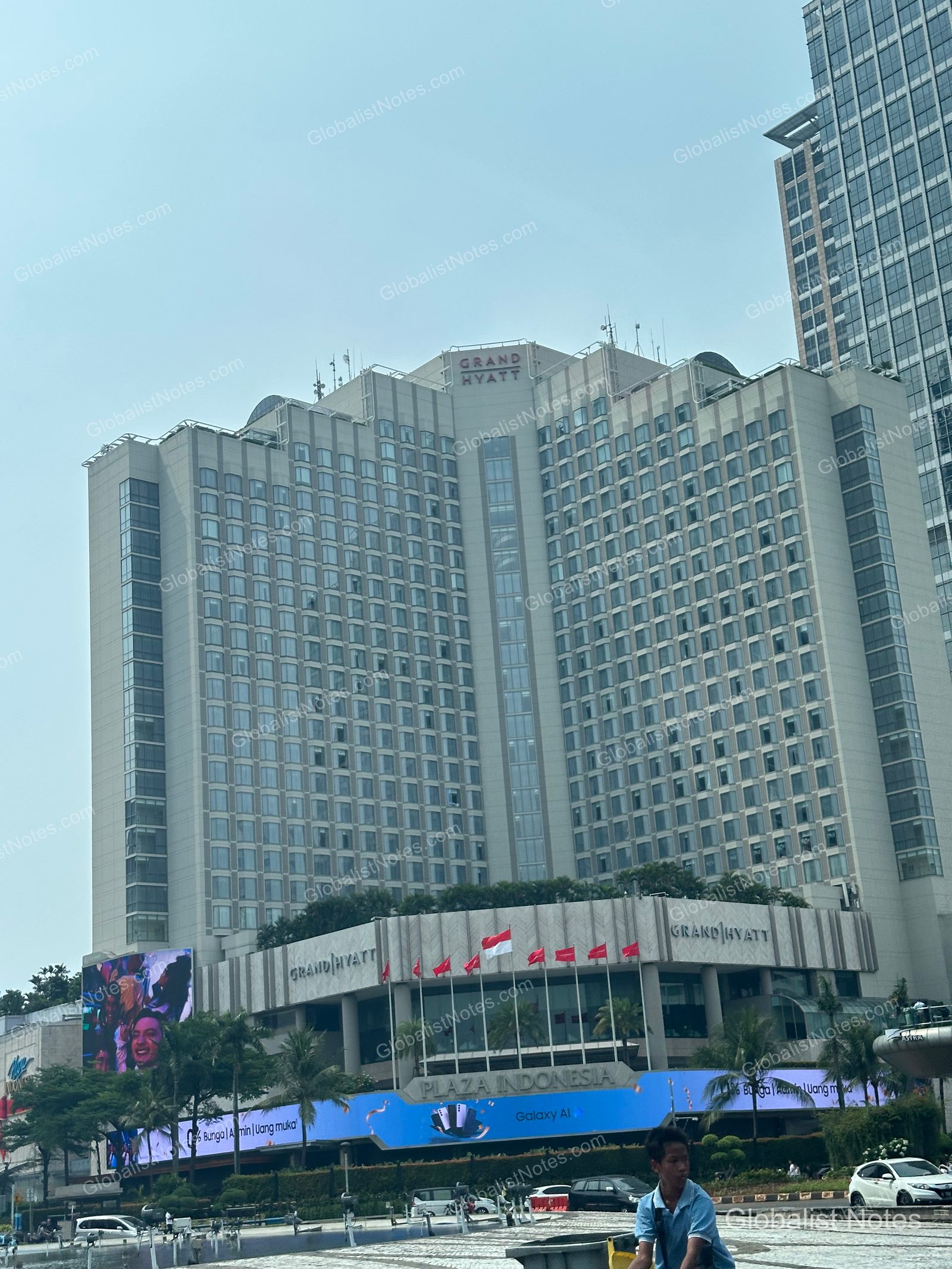 Exterior view of GRAND HYATT Jakarta in the city center. The 'GRAND HYATT' logo is clearly visible on the building facade, with the entrance connecting to the Plaza Indonesia shopping mall and an Indonesian flag flying in front.