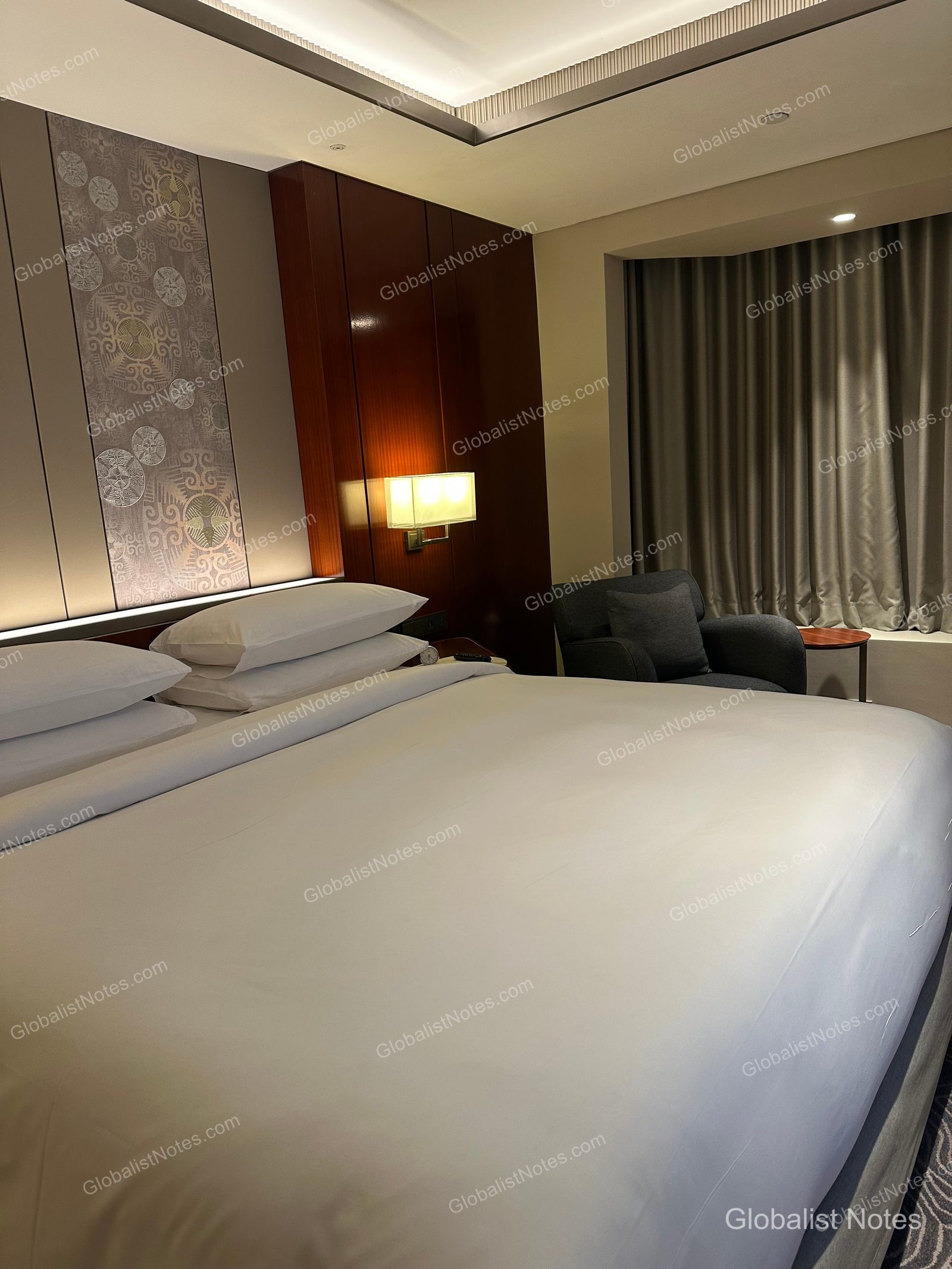 Interior of a Grand Suite at Grand Hyatt Jakarta, featuring a king bed with white linens, wood-paneled walls, soft lighting, and a dark gray armchair with a small round table by the window.