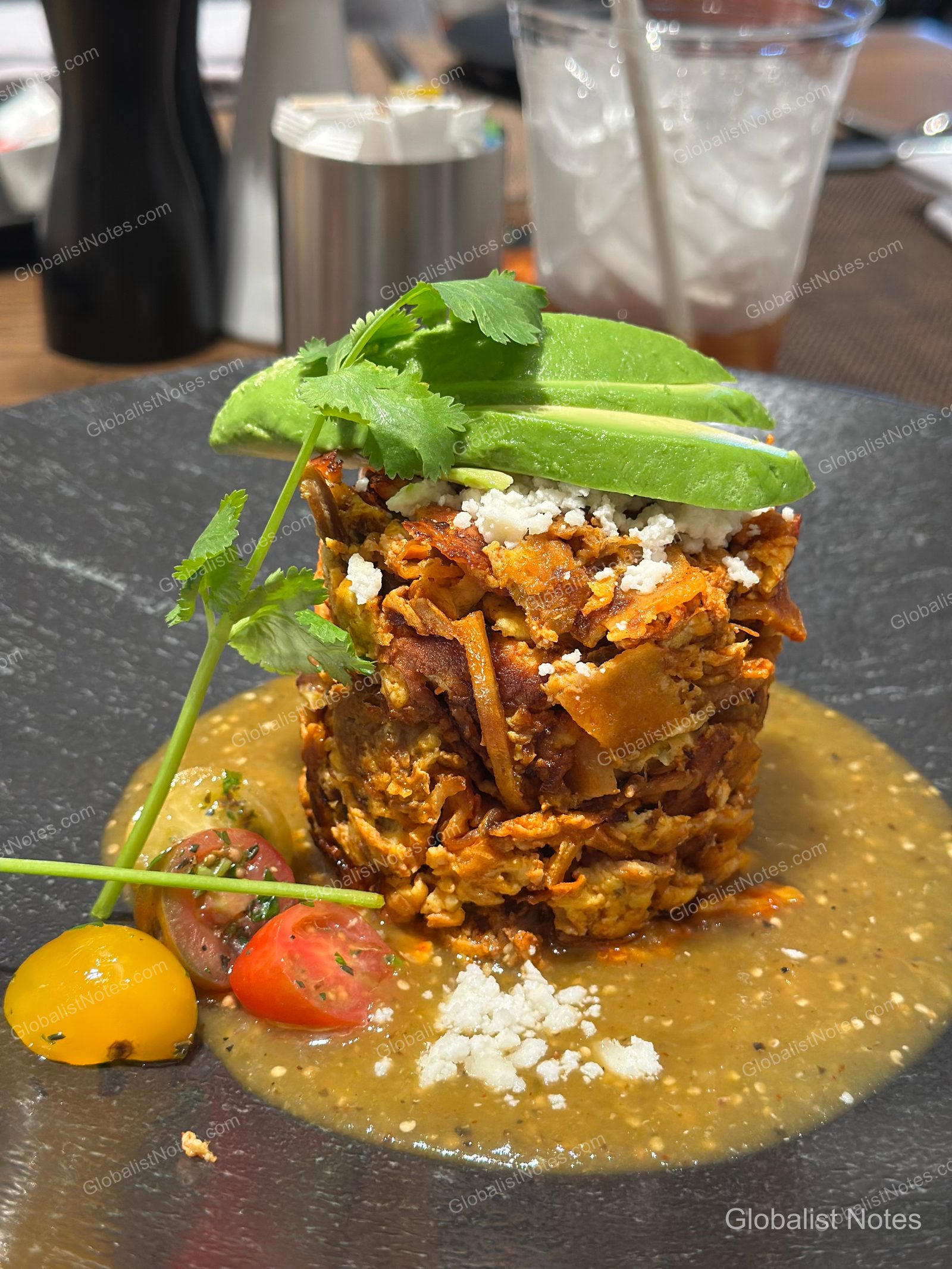 黑色圓盤盛裝的墨西哥玉米片早餐（Chilaquiles），鋪上酪梨切片、香菜與起司粉，並點綴小番茄與綠色醬汁。