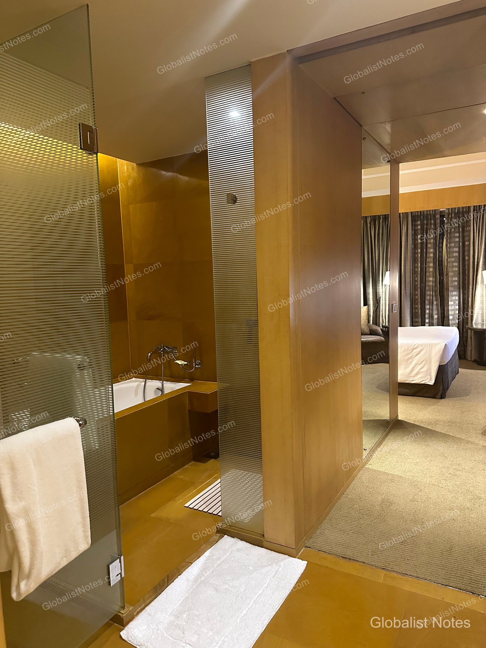 Interior of a Veranda Suite King at Grand Hyatt Mumbai Hotel & Residences. The bathroom, separated by a glass partition, includes a soaking tub and looks into the bedroom, which features warm wood finishes and carpeting.