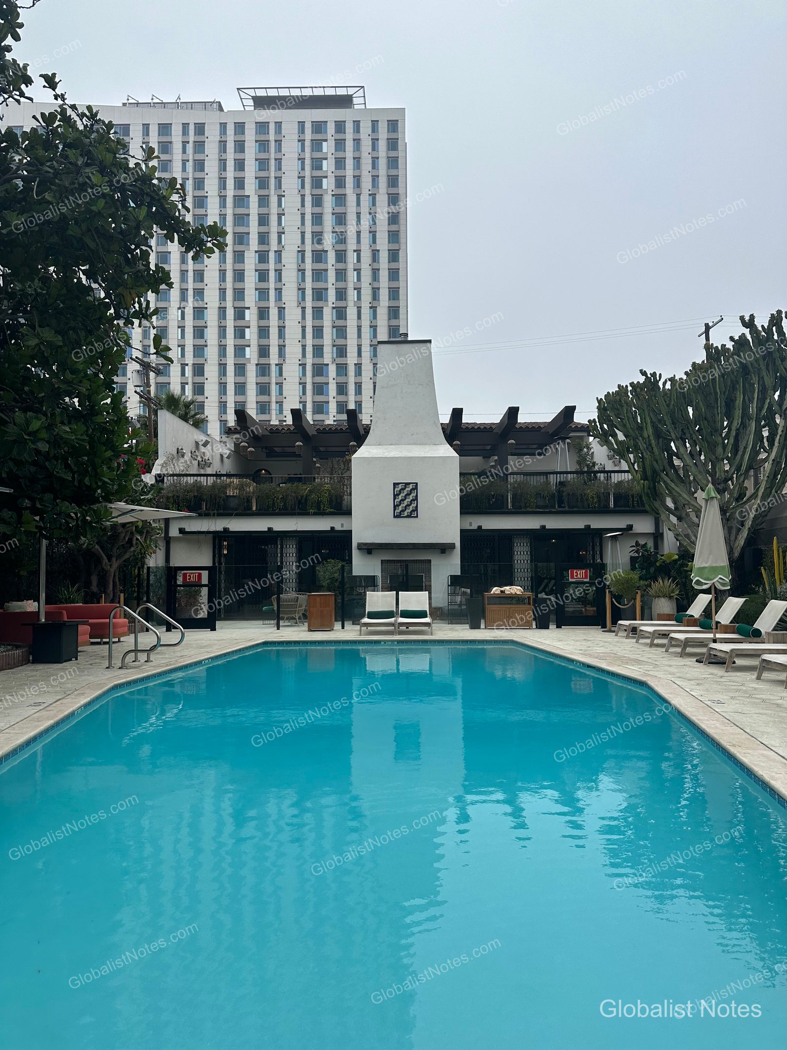 Outdoor pool view at Hotel Figueroa in Los Angeles. A rectangular blue pool is set in front of a large central white fireplace and sun loungers, with high-rise buildings in the background.