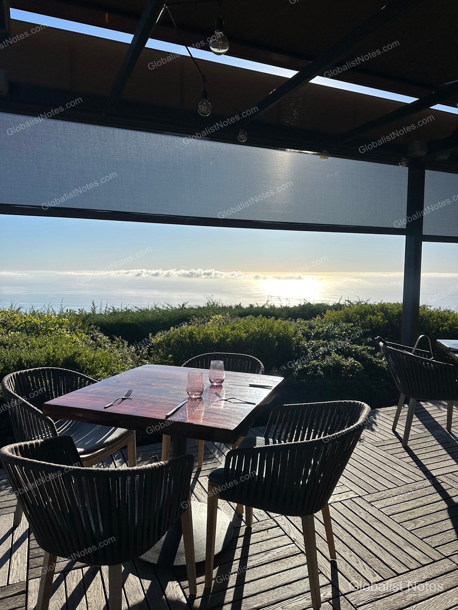 Outdoor terrace dining at Alila Ventana Big Sur, featuring two pink glasses and tableware on a wooden table. The background shows an expansive view of the Pacific Ocean horizon with low-hanging clouds and sunlight reflecting on the water.