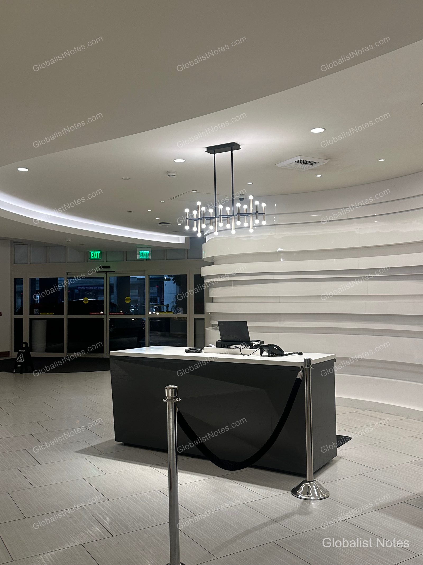 The lobby front desk at the Hyatt Regency near Los Angeles International Airport. Behind the dark-colored desk is a curved white wall, with modern chandelier lighting on the ceiling and light-colored tile flooring.