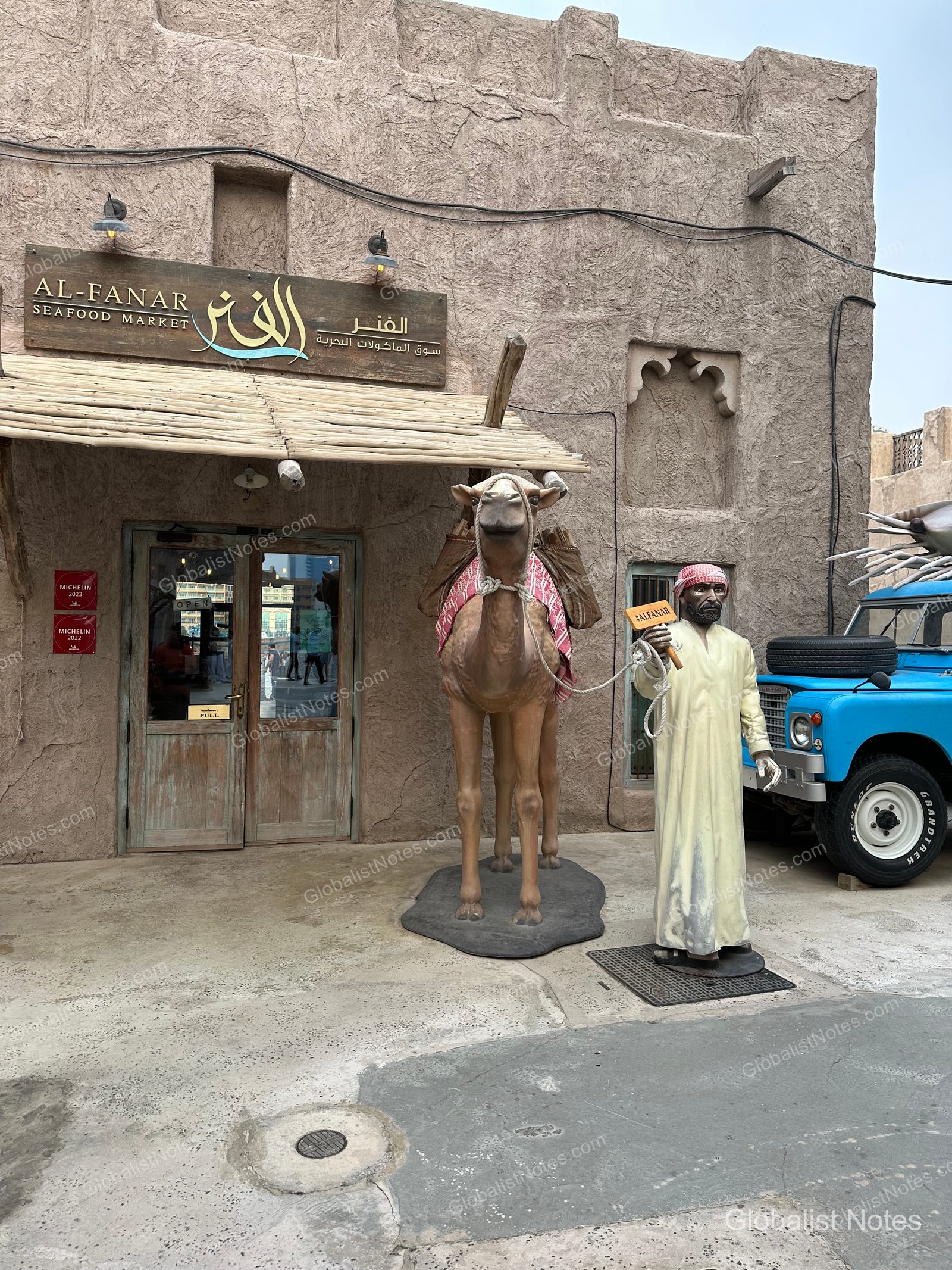 杜拜 Al Fanar Seafood Market 入口，傳統土牆建築前設有駱駝雕像與穿著傳統服飾的人偶，一旁停放著一輛藍色復古汽車。
