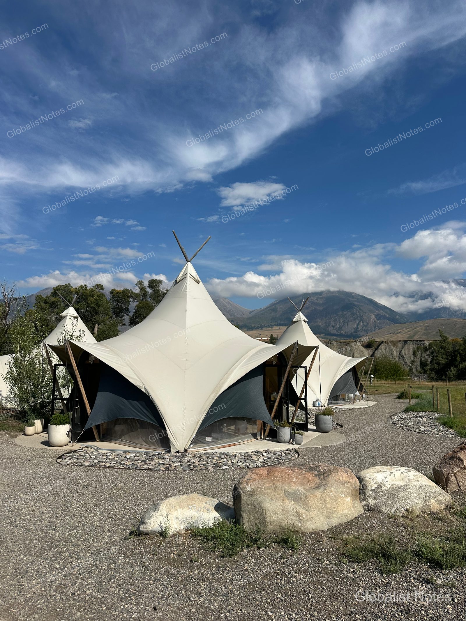 米国パインクリーク、Under Canvas North Yellowstoneのパラダイスバレーに並ぶ大型キャンバステント。砂利道沿いに三角屋根のテントが配置され、背景には澄んだ空とイエローストーン近郊の山々が広がります。