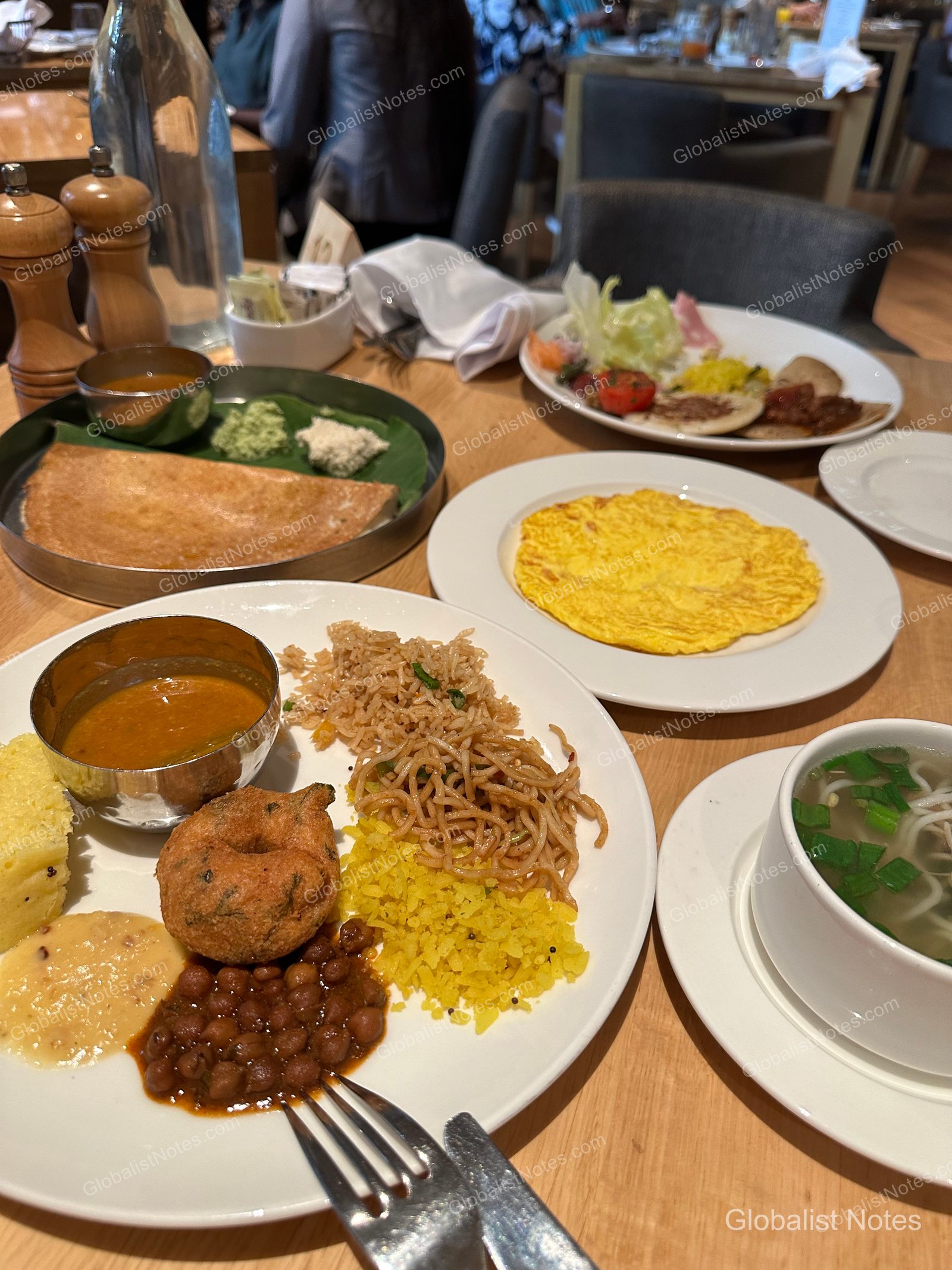 A breakfast buffet spread at Grand Hyatt Mumbai featuring Indian and Asian dishes, including dosa, omelets, stir-fried noodles, poha, vada, and chickpea curry.