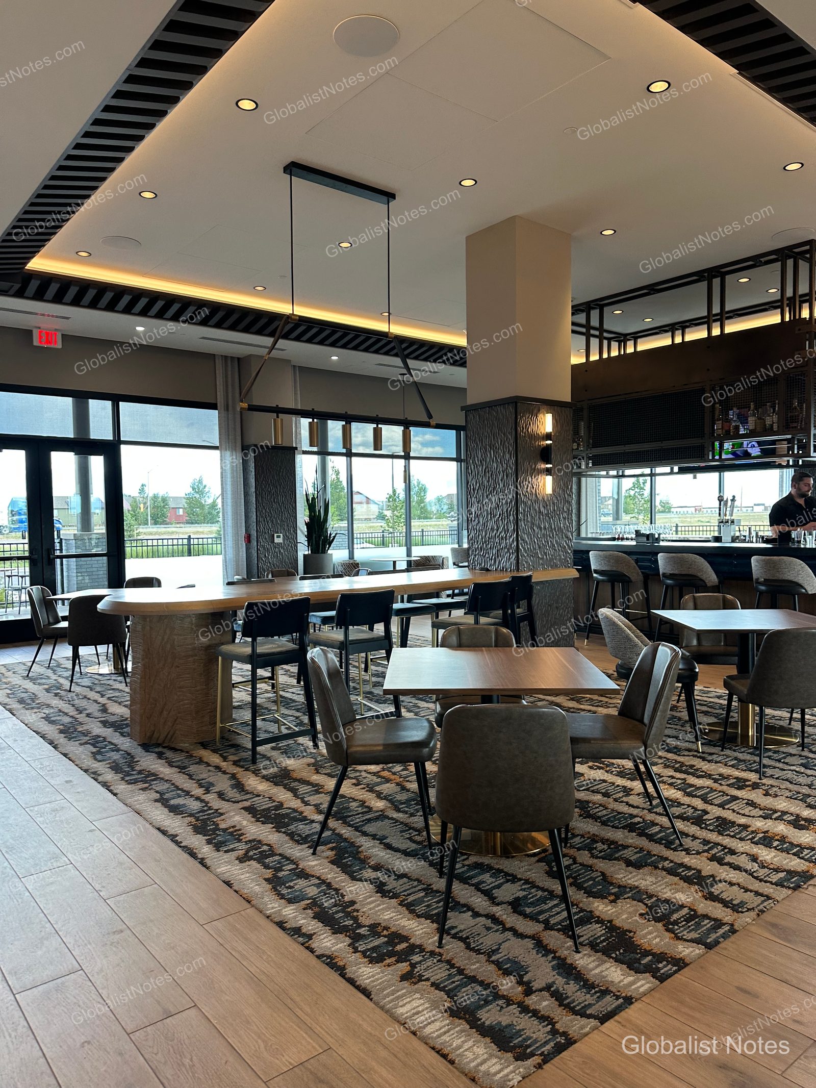 BelgradeにあるHyatt Place Bozeman Yellowstone Airportのロビーバー。長い木製テーブルと椅子が並び、奥にはバーカウンターと外の景色を望む大きな窓があります。床には柄入りのカーペットが敷かれ、天井には間接照明が施されています。