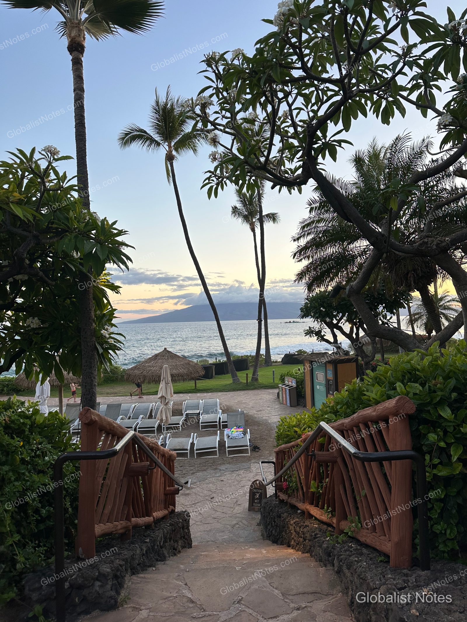 Stone steps leading to the outdoor area at Hyatt Regency Maui Resort and Spa. Wooden railings line both sides of the stairs, with a view of the garden, sun loungers, and the ocean below, set against the silhouette of an island at sunset.