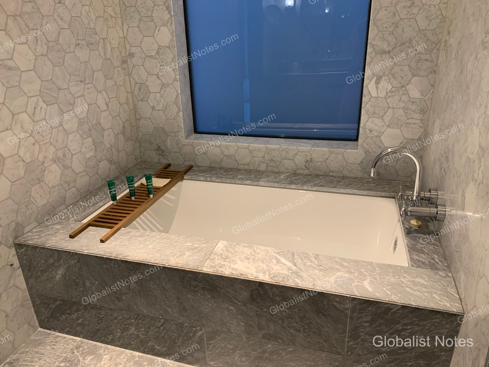 Marble soaking tub in an Alila Napa Valley guest room bathroom, featuring hexagonal wall tiles, a wooden tray across the tub, and a window with a view of the outdoors.