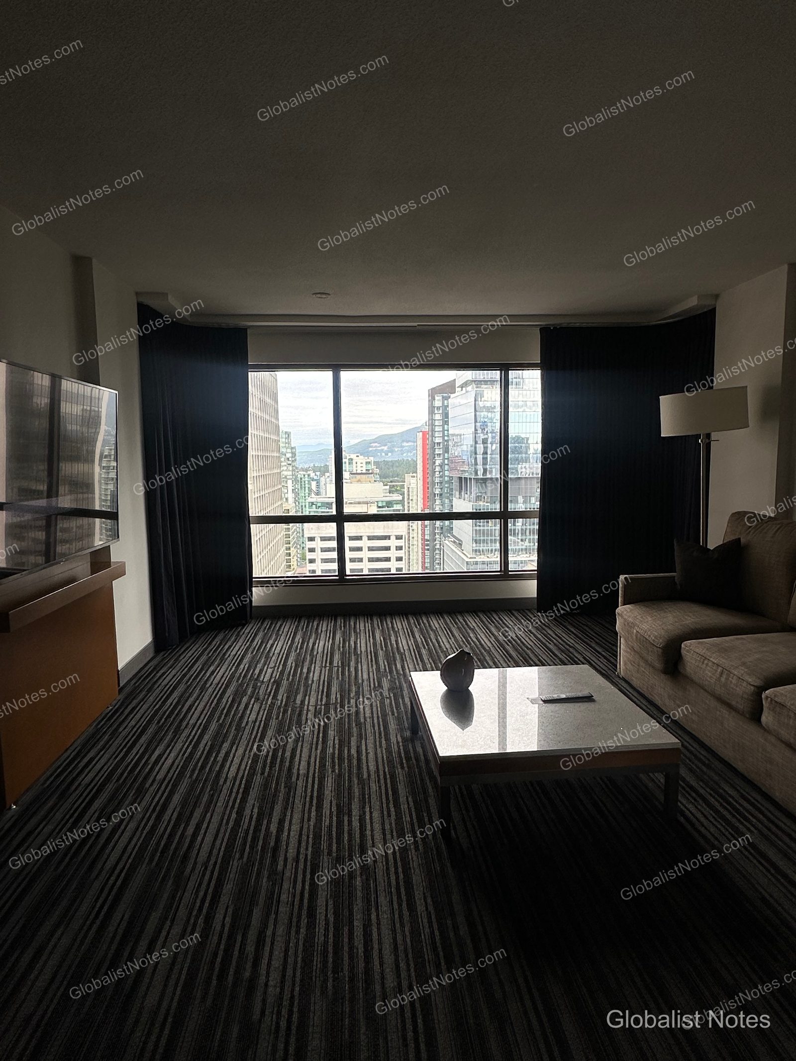Living room view of the Deluxe Suite at Hyatt Regency Vancouver. A glass-topped coffee table and sofa are centered on a striped carpet, with large windows looking out over the downtown Vancouver skyline and distant mountains.