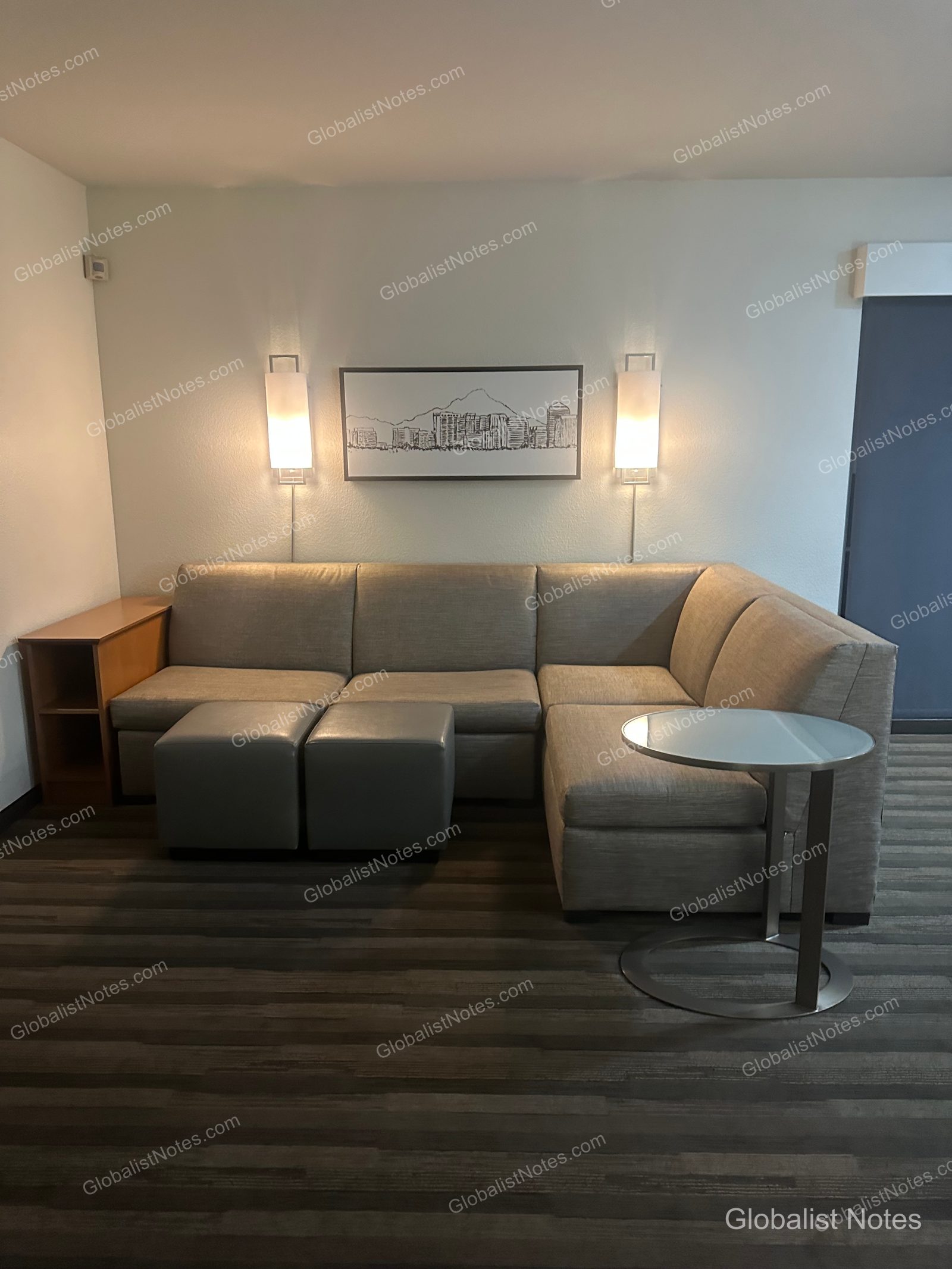 Hyatt House Scottsdale/Old Townの1ベッドルームスイートのリビングエリア。グレーのL字型ソファ、2つのオットマン、円形のサイドテーブルが置かれ、壁にはスコッツデールの街並みを描いたアートとウォールライトが配されている。