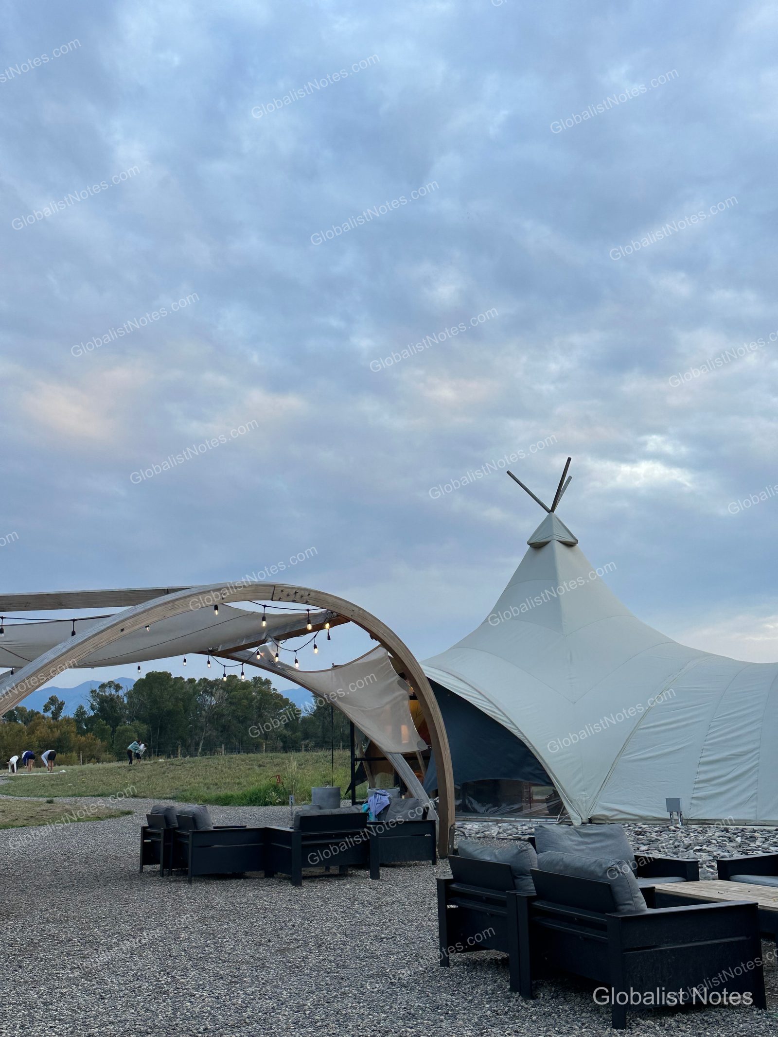 Under Canvas North Yellowstoneの屋外ラウンジ。砂利敷きのスペースに黒いソファと木製フレームの照明が配され、背景には大型テントと曇り空が広がっています。