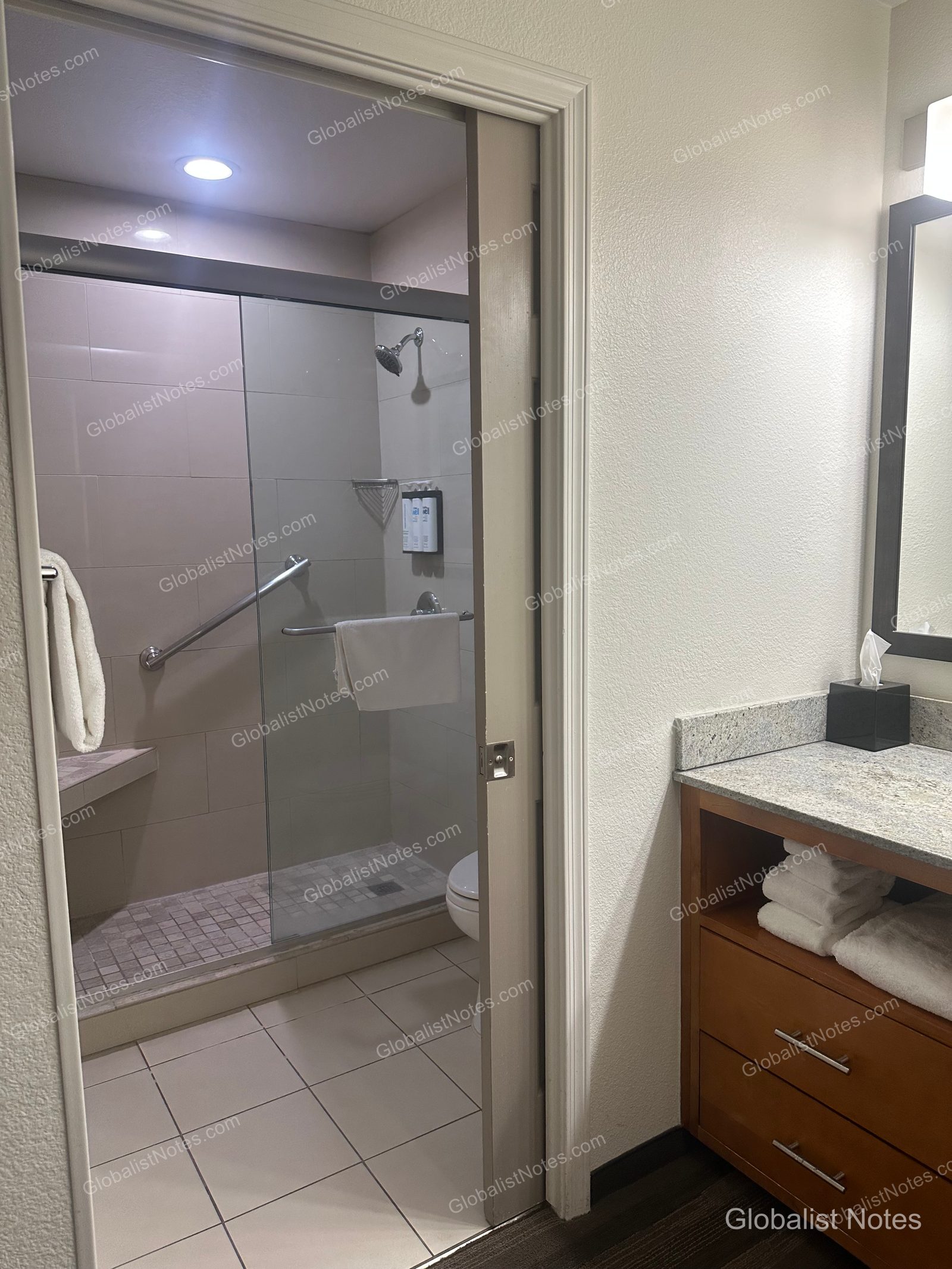 Hyatt House Scottsdale/Old Townのワンベッドルームスイートのバスルーム。ガラス扉のシャワーブース内には手すりとベンチがあり、右手に洗面台が備わっています。