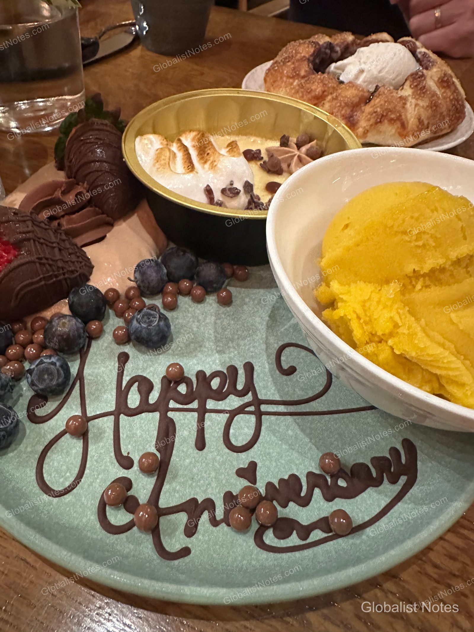 미라발 오스틴 리조트 앤 스파에서 제공된 기념일 디저트 접시입니다. 초콜릿으로 Happy Anniversary 문구가 적힌 민트색 접시 위에 초콜릿 코팅 딸기, 블루베리, 머랭이 올라간 푸딩, 망고 셔벗 볼이 담겨 있으며, 옆에는 페이스트리가 놓여 있습니다.