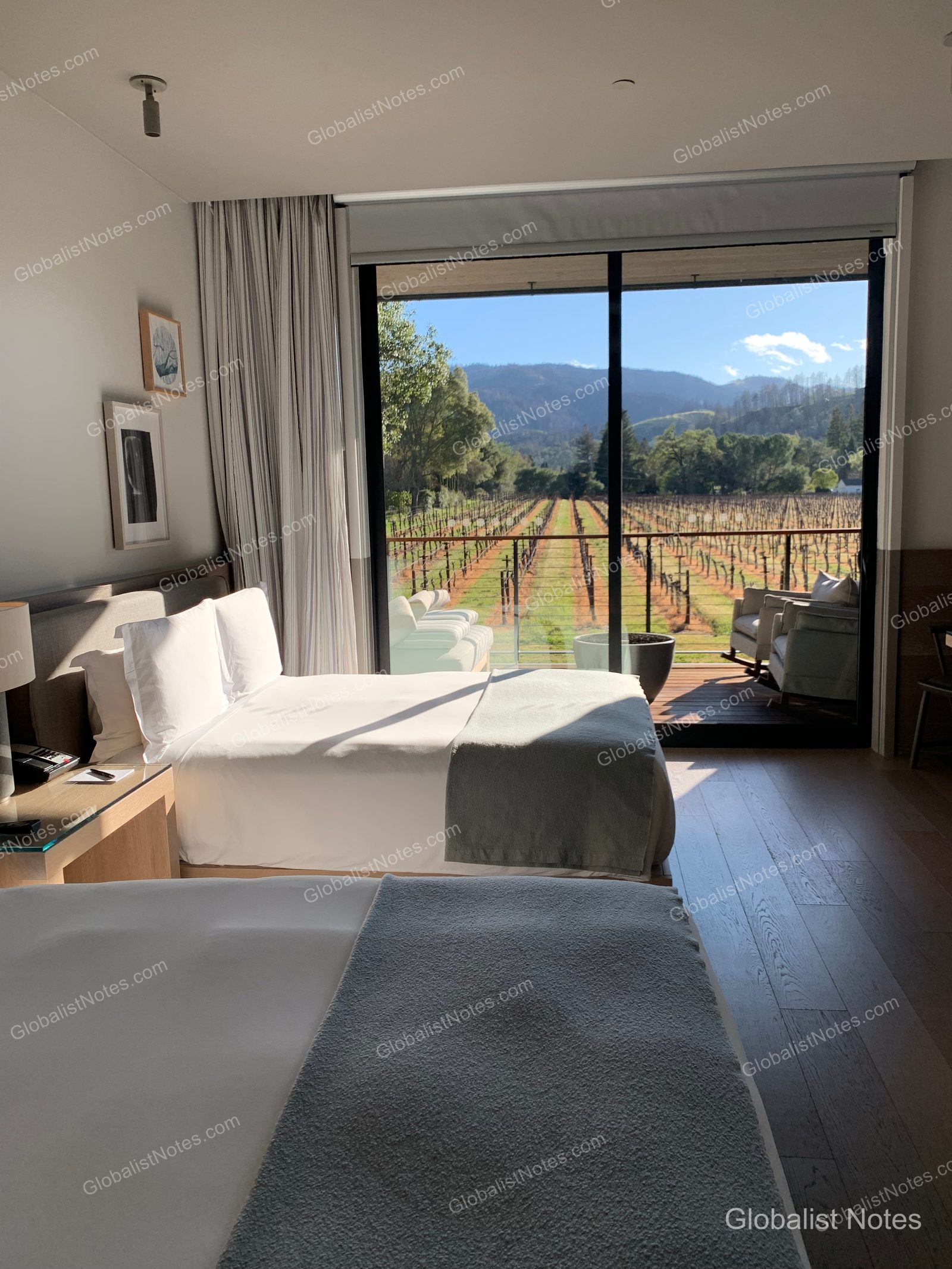 Interior view of a guest room at Alila Napa Valley in St. Helena, USA. A bed with white linens sits next to floor-to-ceiling windows, offering views of the Napa Valley vineyards and distant mountains. The room features wooden flooring and a terrace furnished with chairs.