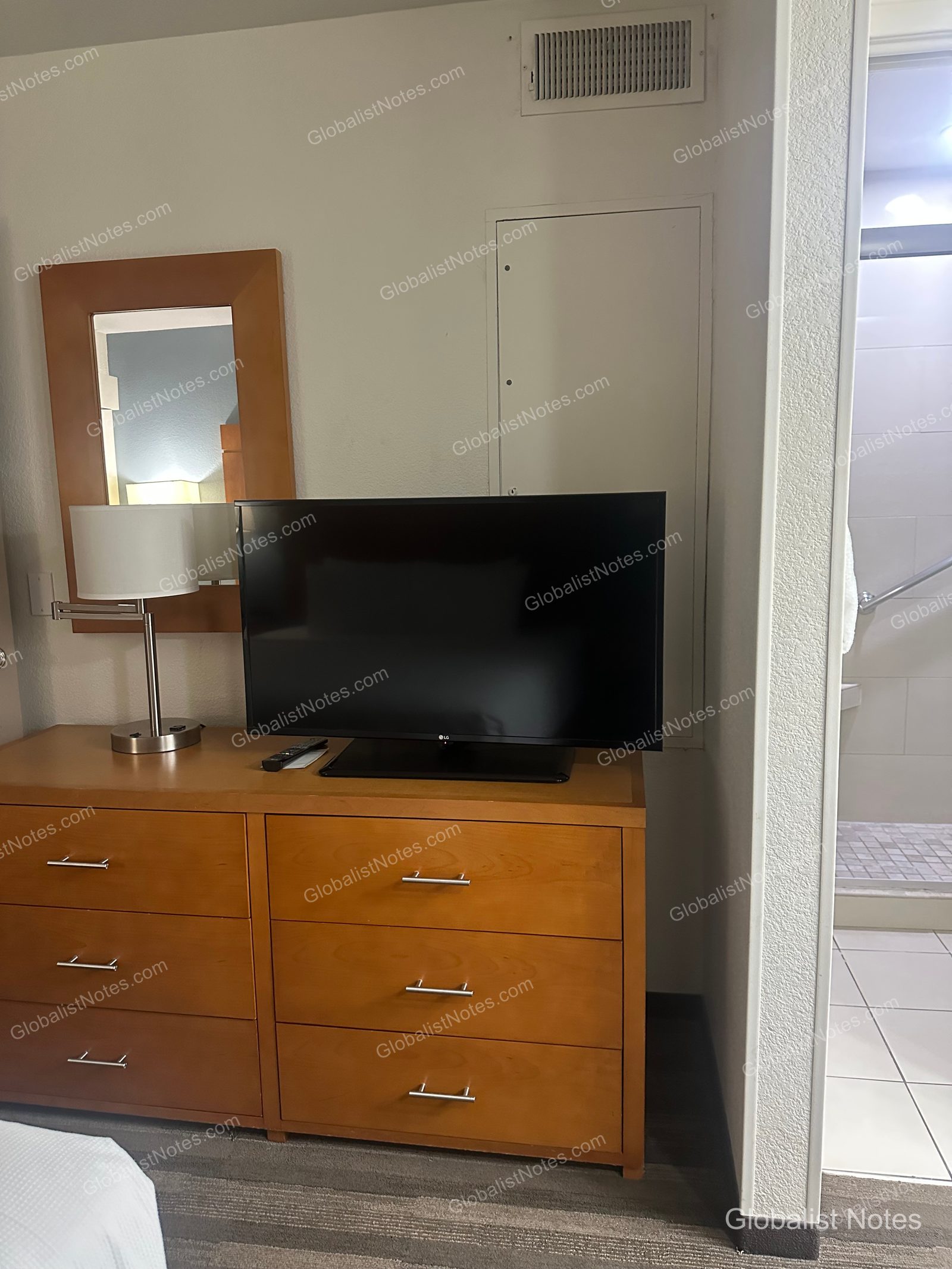 Hyatt House Scottsdale/Old Townのワンベッドルームスイート。木製チェストの上にテレビとスタンドライトが置かれ、右手にバスルームの入り口が見えます。