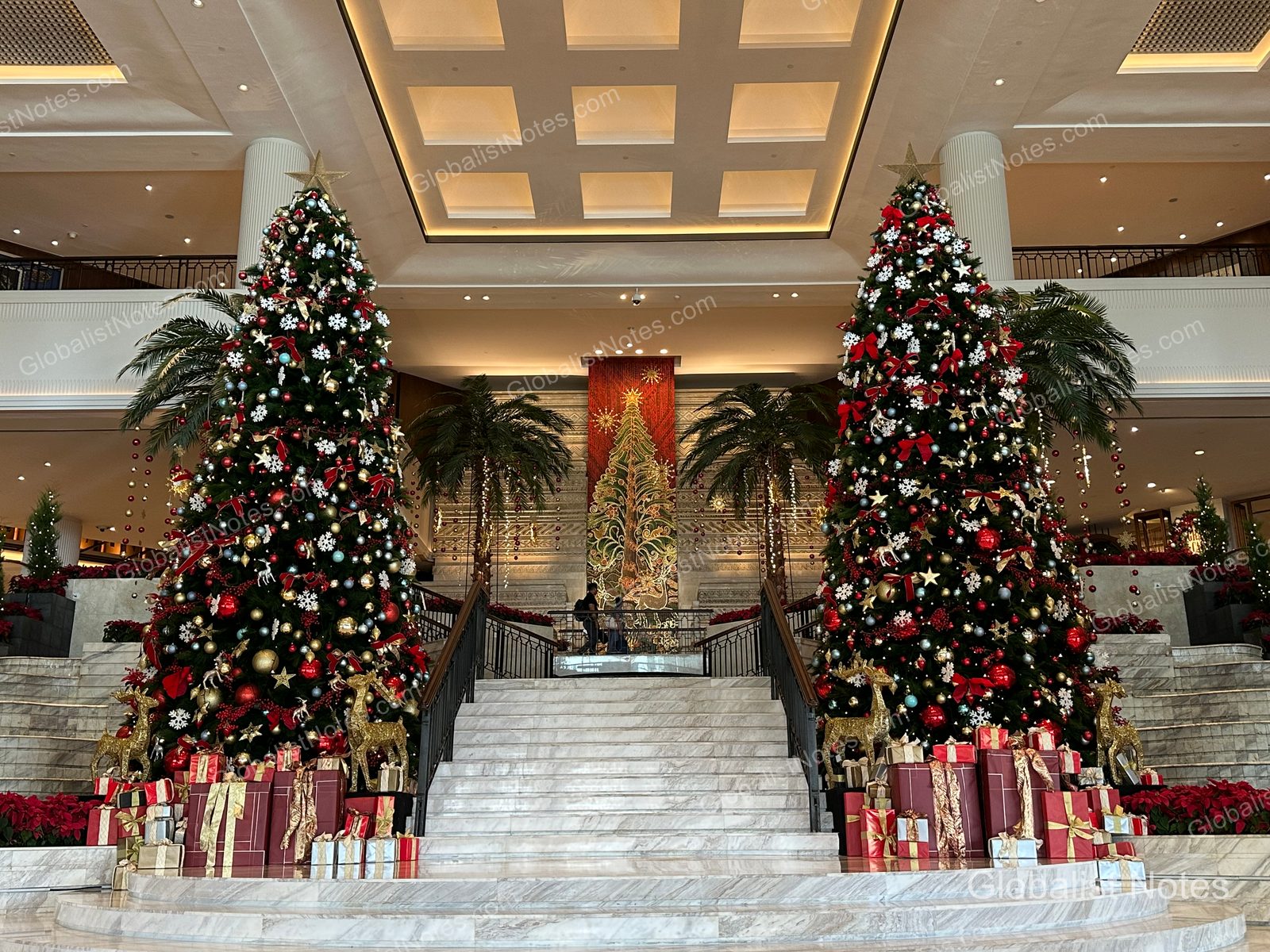 Two large Christmas trees decorated in red and gold stand on either side of the marble staircase in the Grand Hyatt Jakarta lobby, with gift boxes arranged at the base of the stairs.