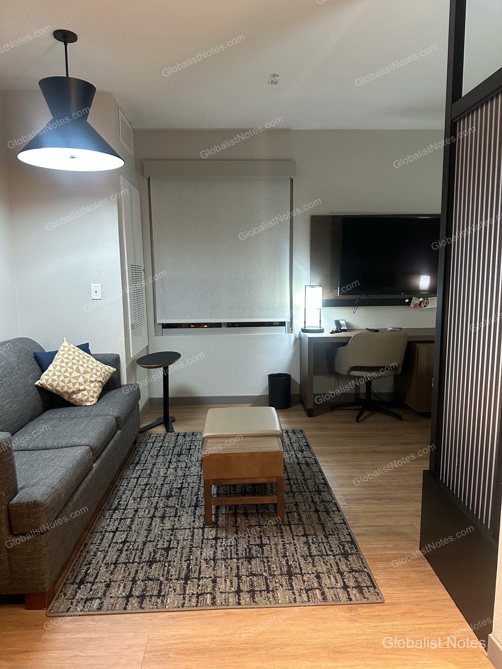 Hyatt Place Flagstaffのデラックスルームのリビングエリア。グレーのソファ、柄物のクッション、小テーブル、オットマン、壁掛けテレビ、ワークデスクが配され、床には幾何学模様のラグが敷かれています。