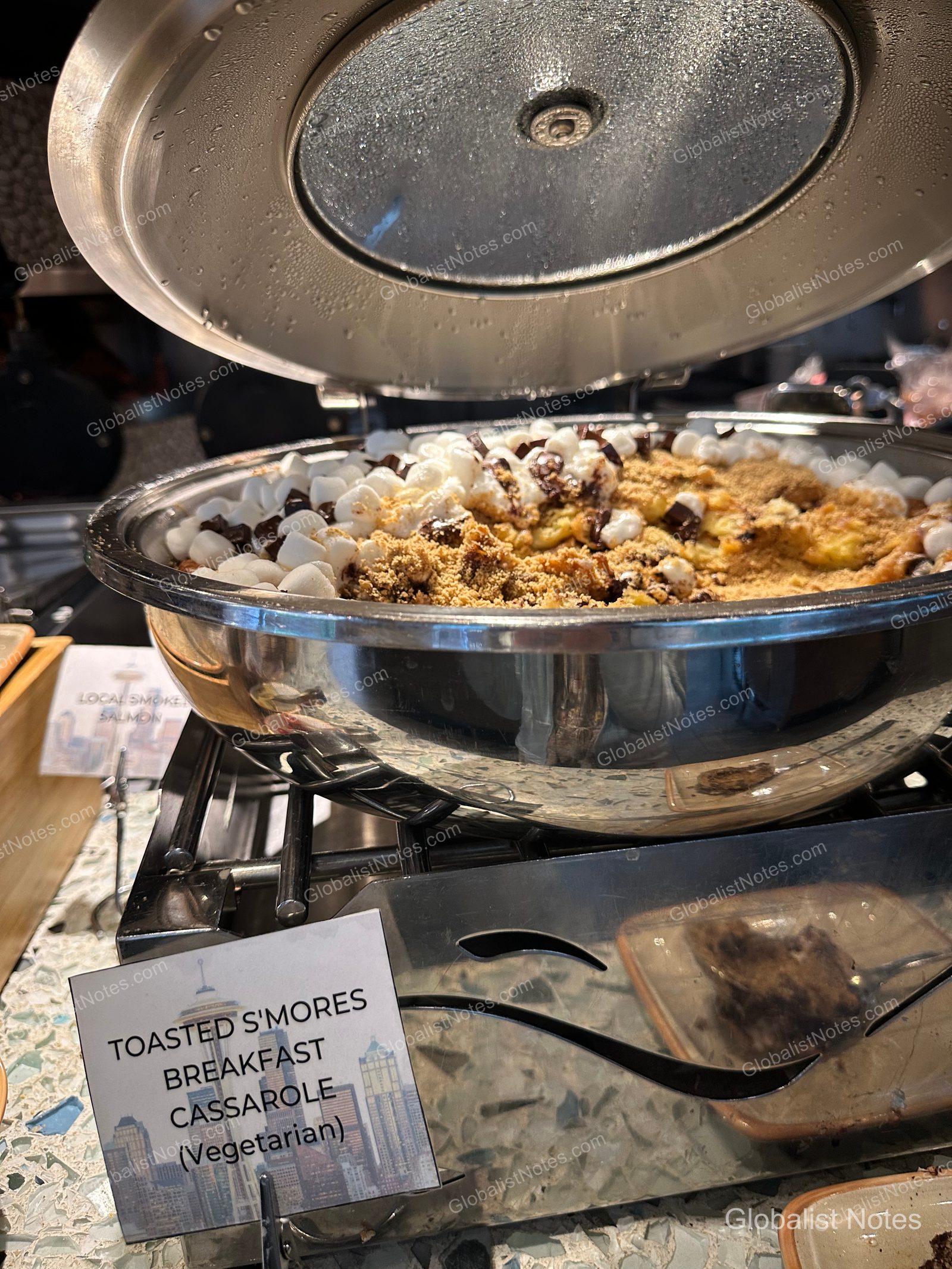 Toasted s'mores breakfast casserole in a silver warmer at the Hyatt Regency Seattle breakfast buffet. The warm dish is topped with marshmallows and graham cracker crumbs, with a menu card in front indicating the dish name and vegetarian status.