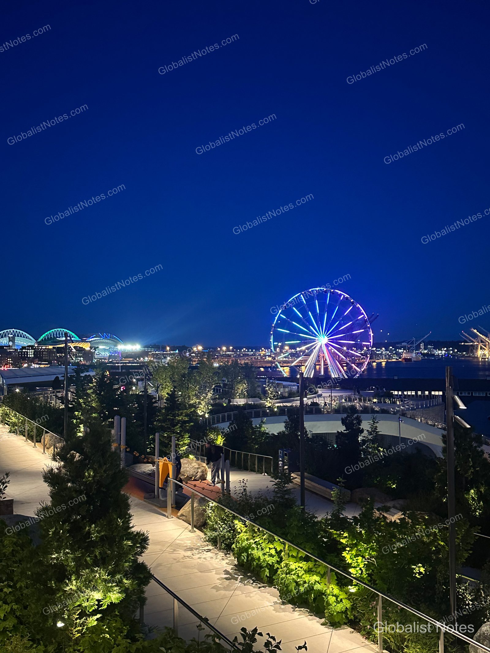 The Seattle Great Wheel and waterfront promenade illuminated at night. The stadium lights are visible on the left, with the harbor lights creating a view of the downtown Seattle skyline.
