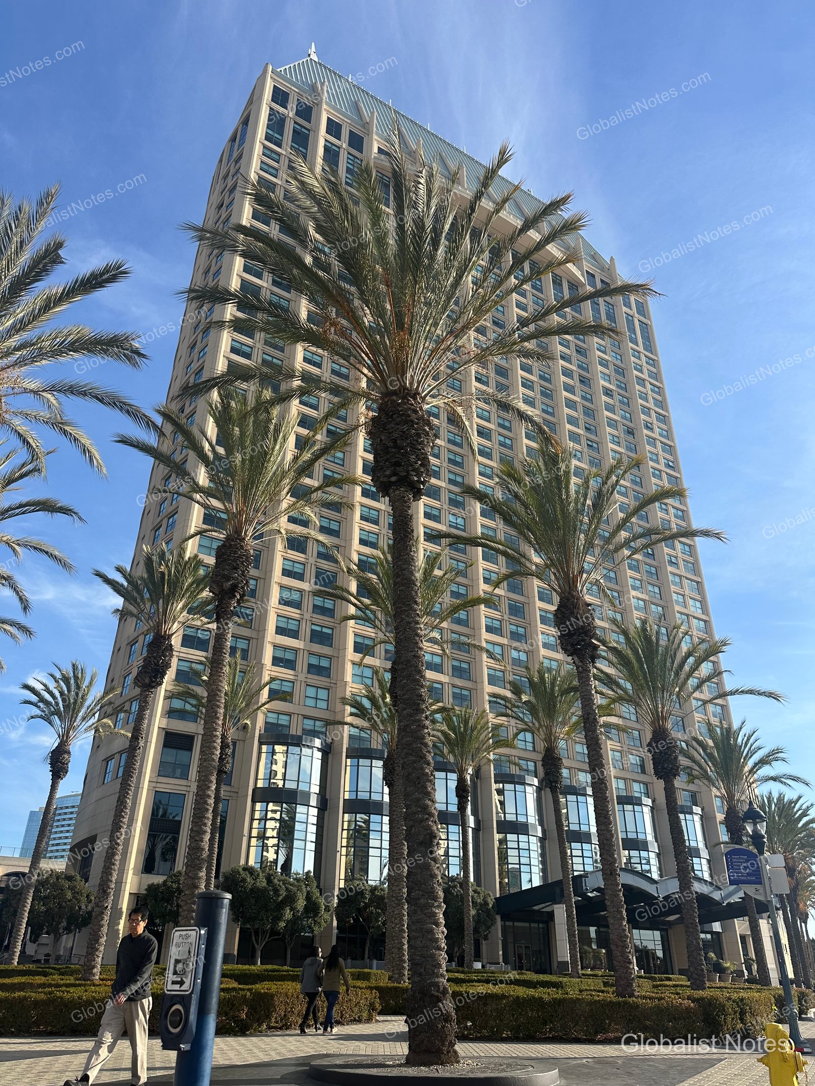 Manchester Grand Hyatt San Diego 高聳的建築外觀在聖地牙哥的晴空下十分顯眼。飯店前方種植著高大的棕櫚樹，人行道上有行人經過，並可見通往入口的景觀步道。