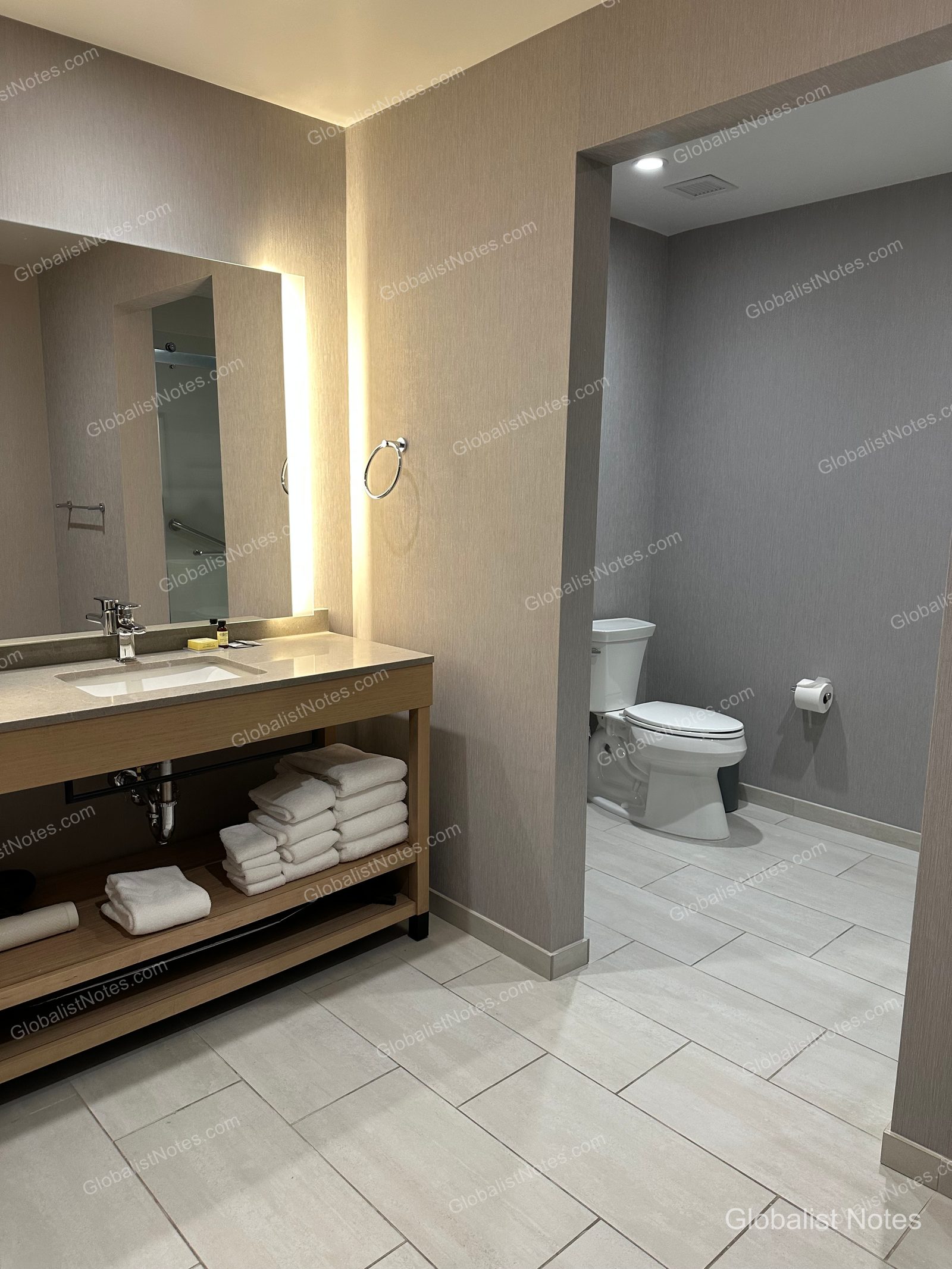 Hyatt Place Bozeman Yellowstone Airportのジュニアスイートのバスルーム。左側に照明付きの鏡と洗面台、下段の棚にタオルが整えられており、右側にトイレが配置されています。