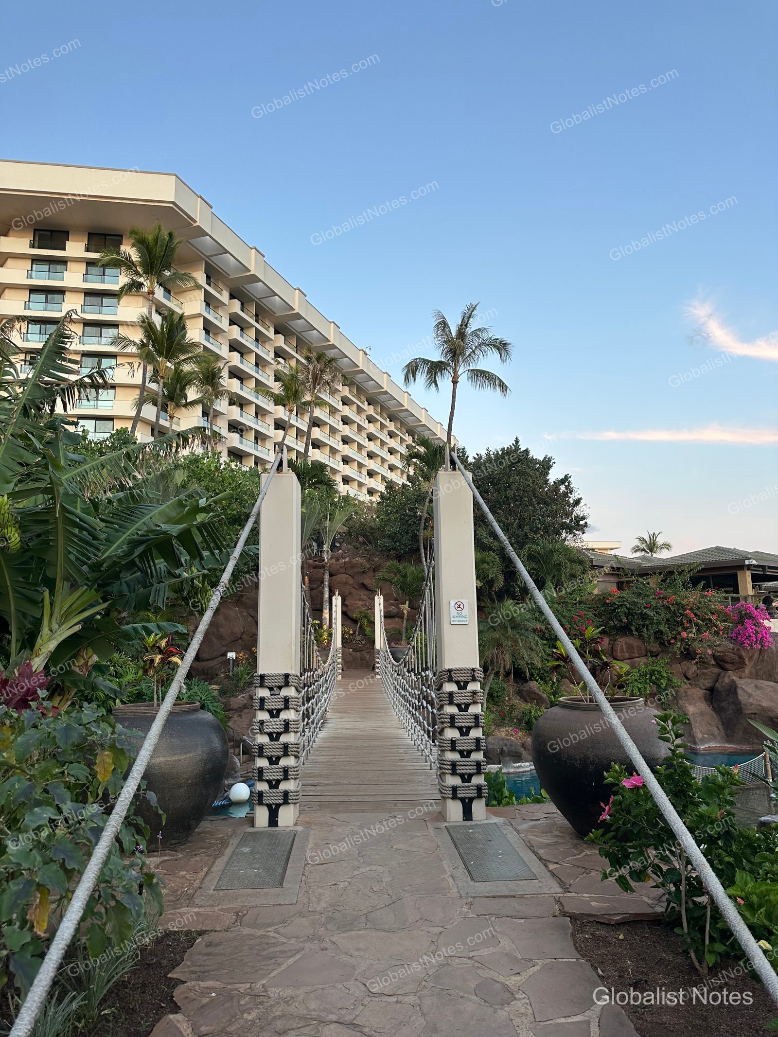 Outdoor suspension bridge at Hyatt Regency Maui Resort and Spa in Lahaina, Hawaii. The bridge, featuring wooden decking and rope railings, connects the resort buildings through tropical gardens surrounded by palm trees and potted plants.