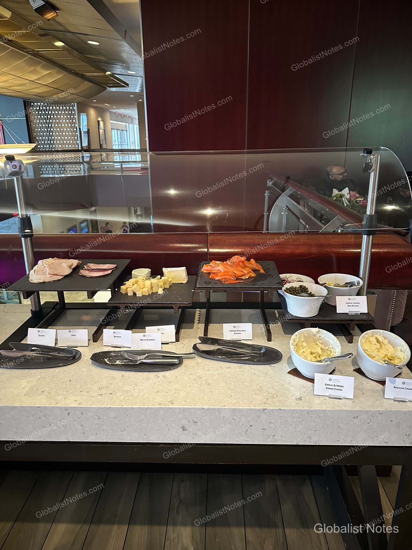 Cold section of the Hyatt Regency Vancouver breakfast buffet featuring smoked salmon, ham, salami, and assorted cheeses on black slate platters, accompanied by capers, red onion, and two types of cream cheese in white bowls, all protected by a clear sneeze guard.