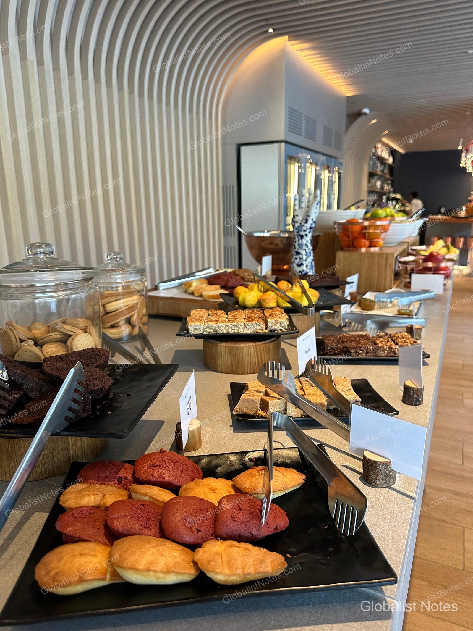 The dessert station at the Hyatt Regency Malta breakfast buffet, with madeleines, brownies, and sliced cakes on black square plates, plus cookies in glass jars. A curved wall and bowls of fruit are visible in the background.