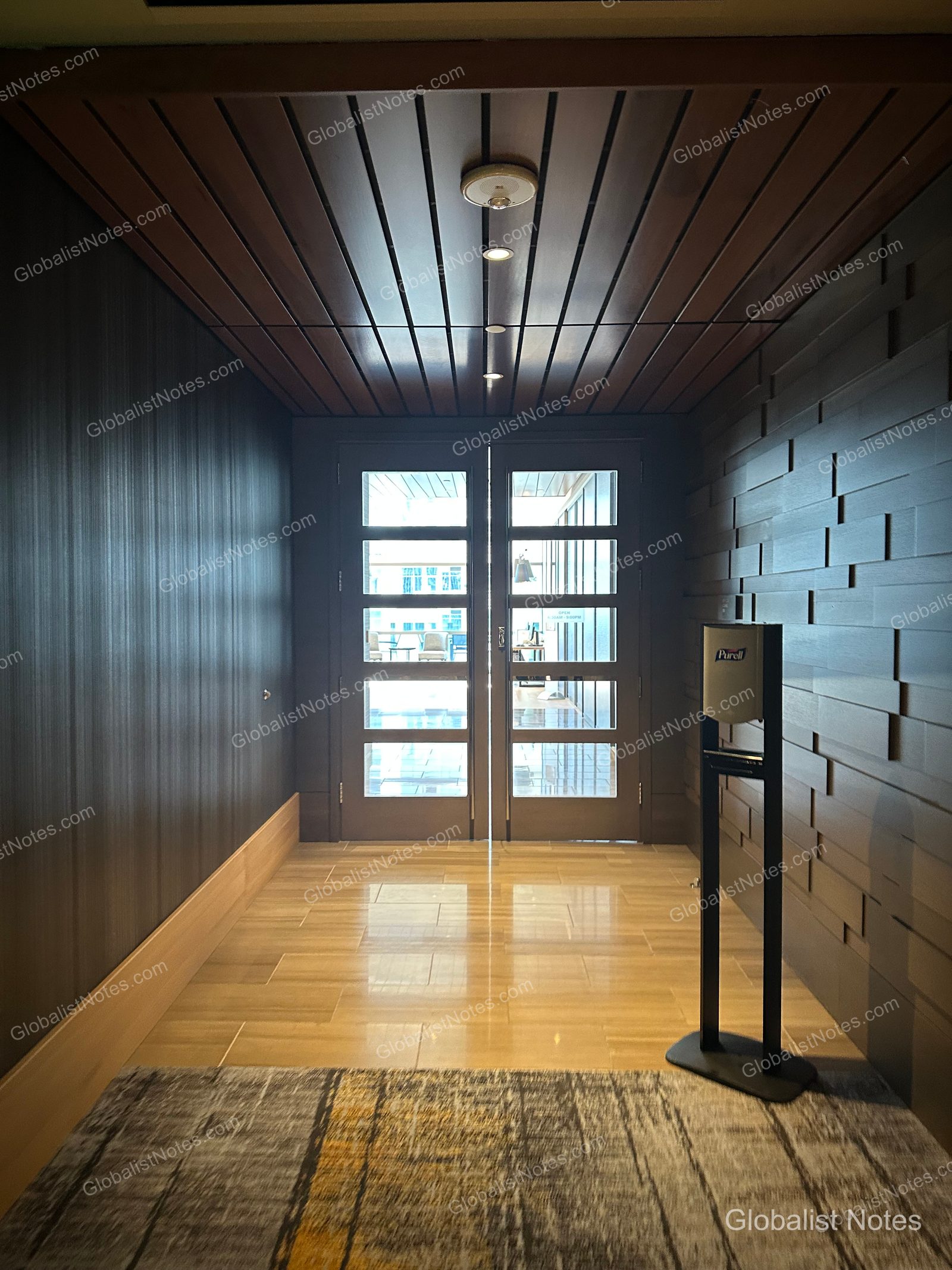 A hallway at Hyatt Regency Seattle featuring dark wood paneling, ending in a set of double glass doors. A black freestanding hand sanitizer dispenser stands on the right. The floor is a mix of light-colored tile and patterned carpet.