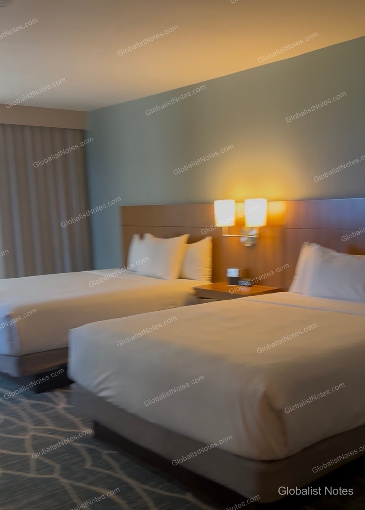 Interior view of a Partial Ocean View room at Hyatt Place Waikiki Beach in Honolulu, Hawaii. Two beds are placed side-by-side, featuring a wood-tone headboard with integrated lighting and a small nightstand between them. The floor is finished with patterned carpeting.