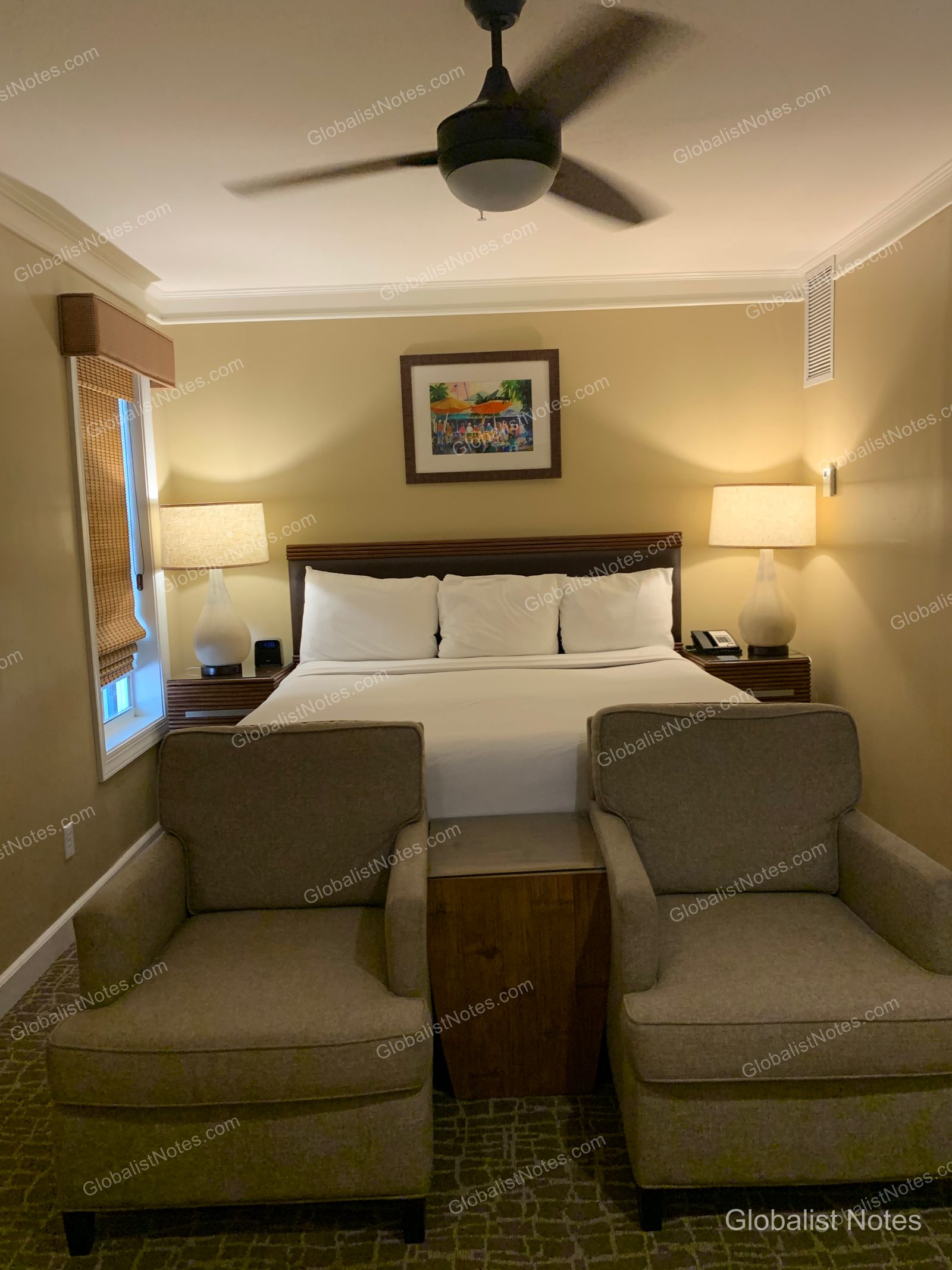 Hyatt Vacation Club at Sunset Harborの客室。中央に白い寝具のキングベッドが配され、両サイドにサイドテーブルと照明が備わっています。ベッドの足元にはアームチェア2脚と小さなテーブルがあり、天井にはシーリングファンが設置されています。