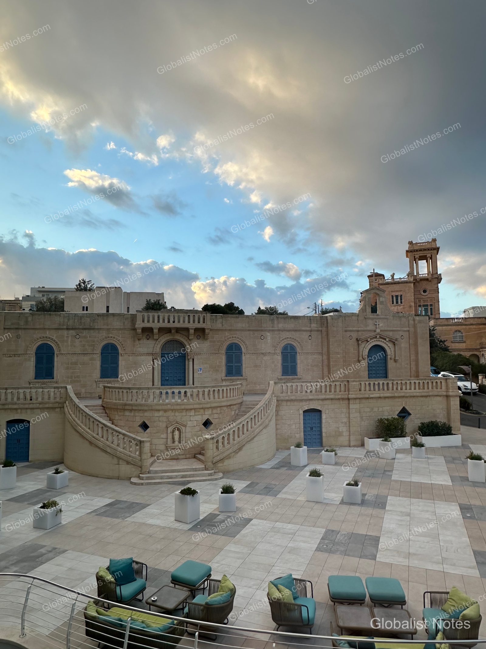 The outdoor terrace at Hyatt Regency Malta in St. Julian's, where modern furniture sits against a backdrop of historic stone architecture and blue doors under a cloudy sunset sky.