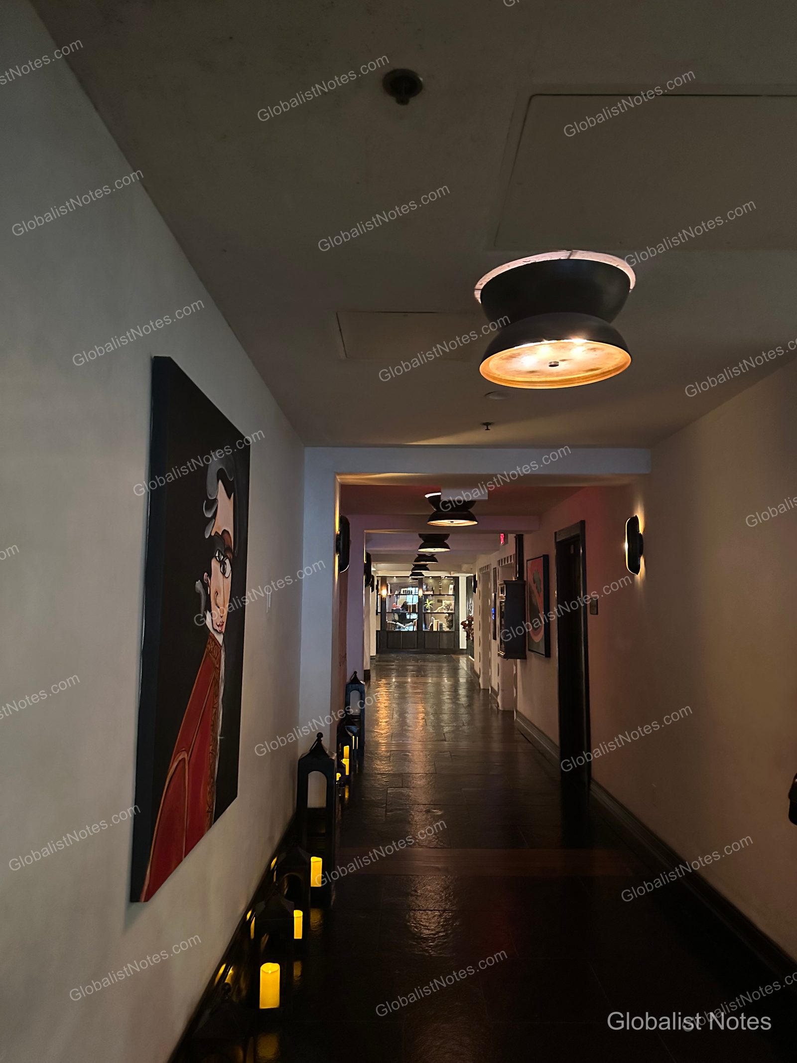 A dimly lit hallway at Hotel Figueroa in Los Angeles, featuring portraits on the walls, a row of lanterns along the floor, and a series of circular ceiling lights extending down the corridor.