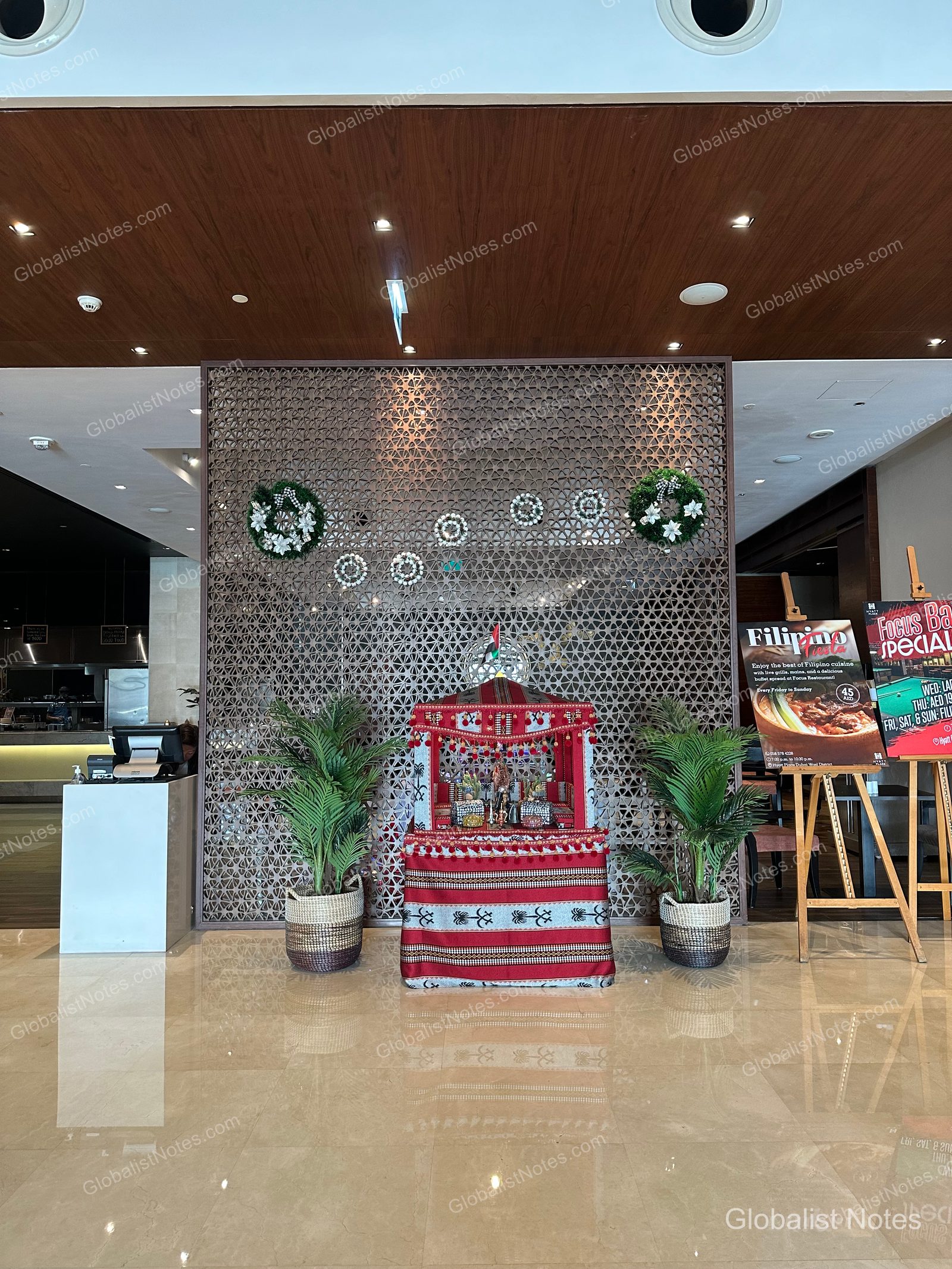 Hyatt Place Dubai/Wasl District 大廳中央設有一面阿拉伯風格裝飾牆，前方擺放著鋪設紅色傳統布料的展示台與兩盆植栽。牆上掛著聖誕花圈，一旁則立著餐廳的促銷立牌。