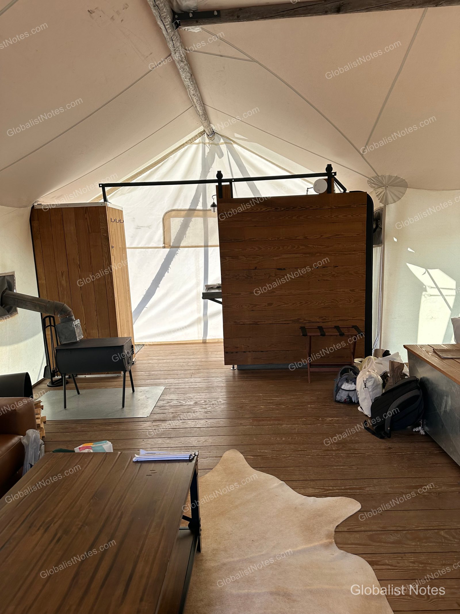 Under Canvas North Yellowstoneのジュニアスイートテント内部。木製の床に薪ストーブとレザーラグが置かれ、中央にはバスルームを仕切る木製パーティションがあります。キャンバス地の天井から自然光が差し込んでいます。