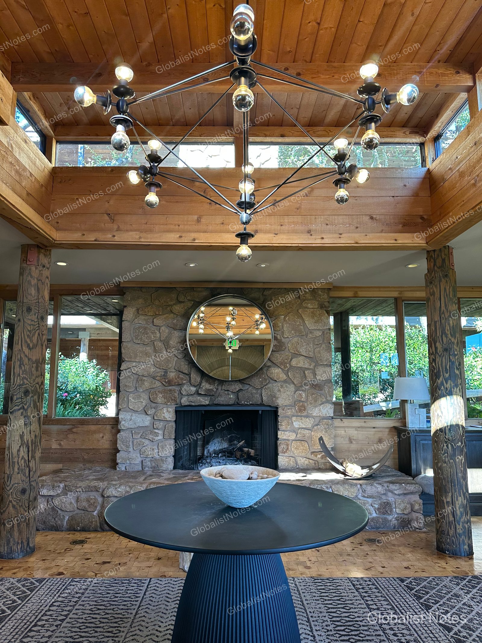 Lobby area at Alila Ventana Big Sur featuring high wood-paneled ceilings with geometric chandeliers, a central stone fireplace, and a circular table. Floor-to-ceiling windows provide natural light, highlighting the warm, neutral wood-toned interior.