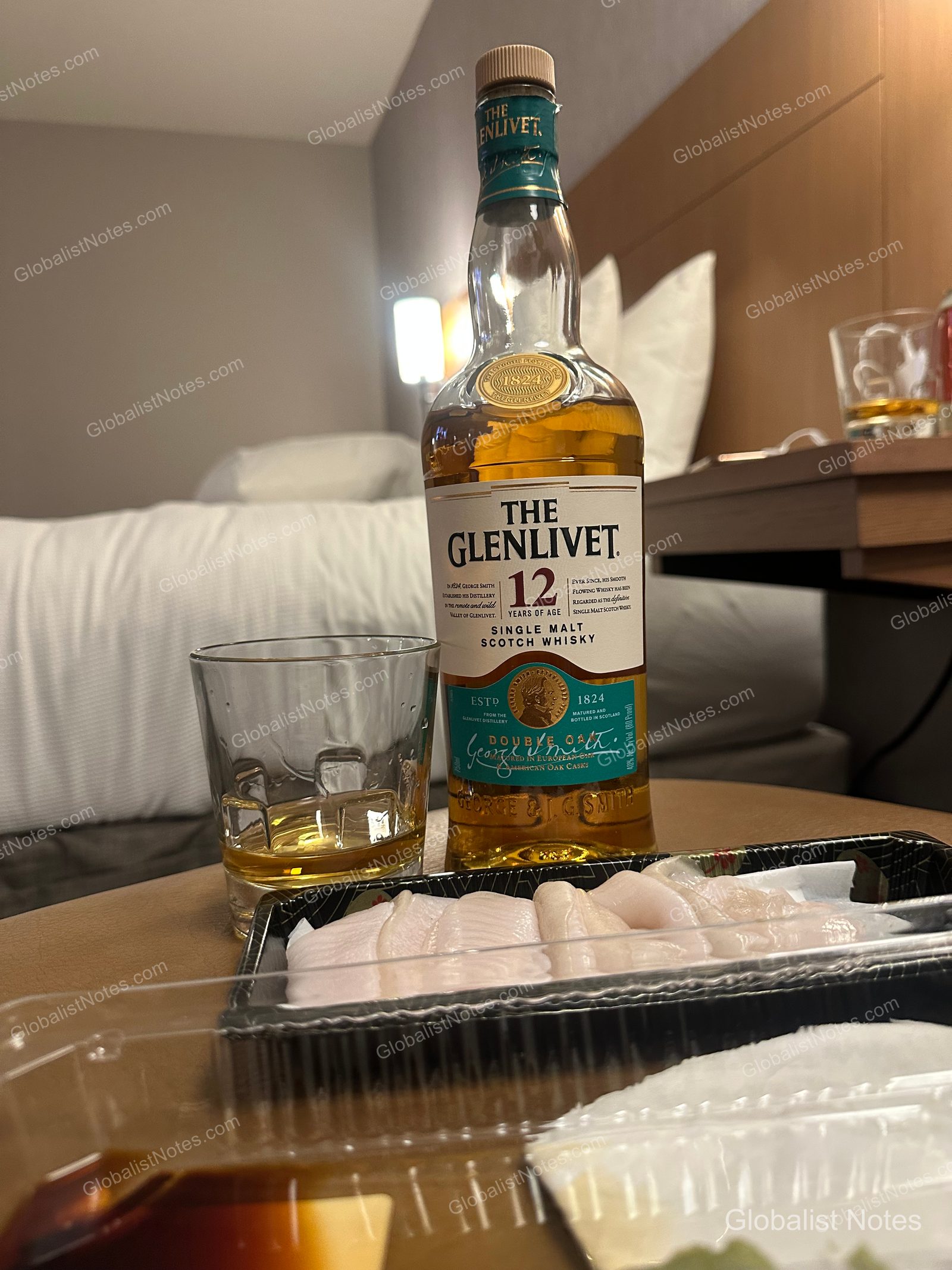 Hyatt Place Tucson-Central 初級套房內，桌上擺放著一瓶 The Glenlivet 12 年威士忌、酒杯及外帶生魚片，背景為模糊的飯店床鋪。