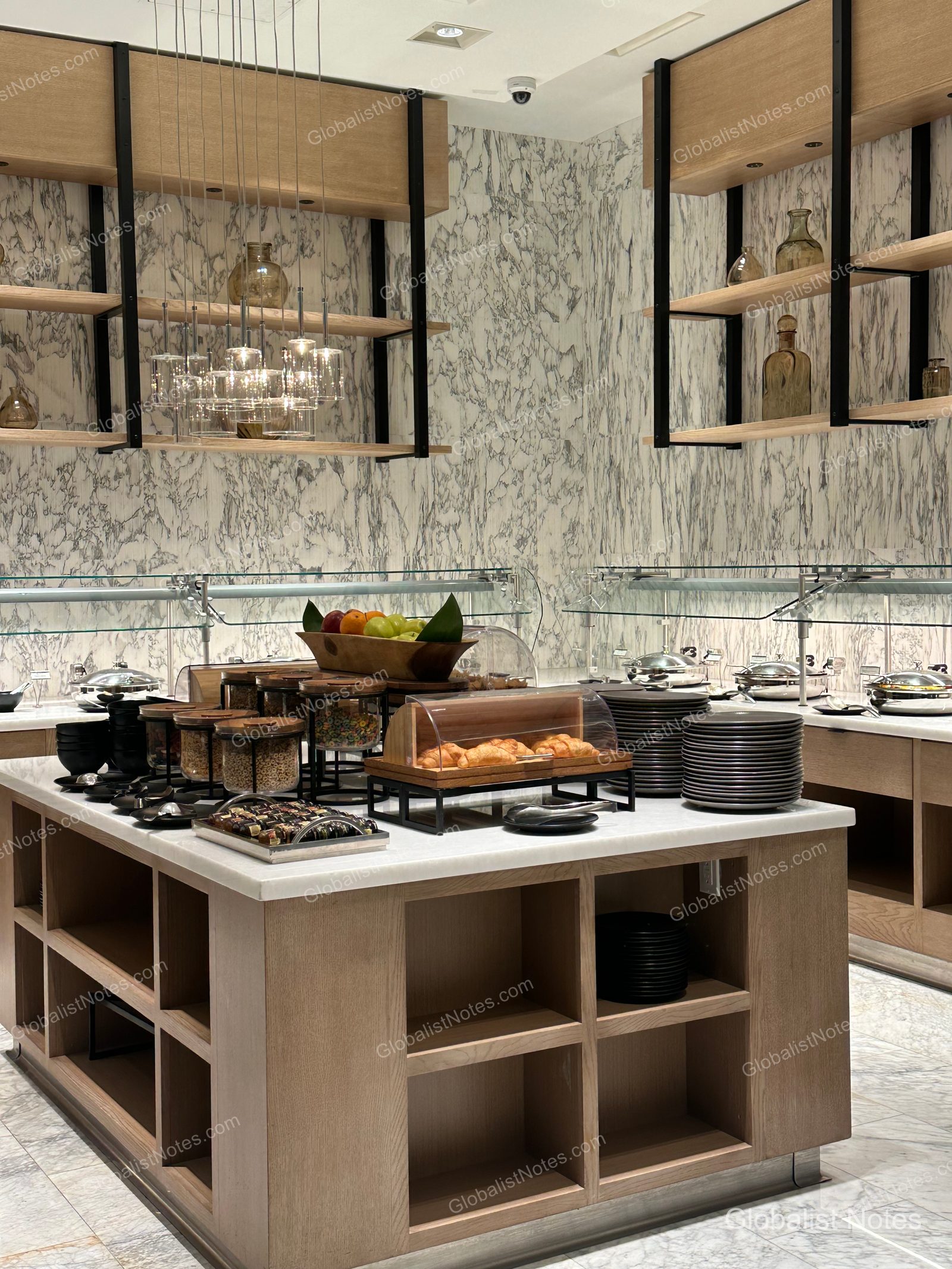 Hyatt Regency Newport Beach West 早餐自助餐區，大理石牆面搭配木質層架與燈光。中央島檯整齊擺放著可頌、水果、麥片與餐盤，後方則為熱食保溫鍋。