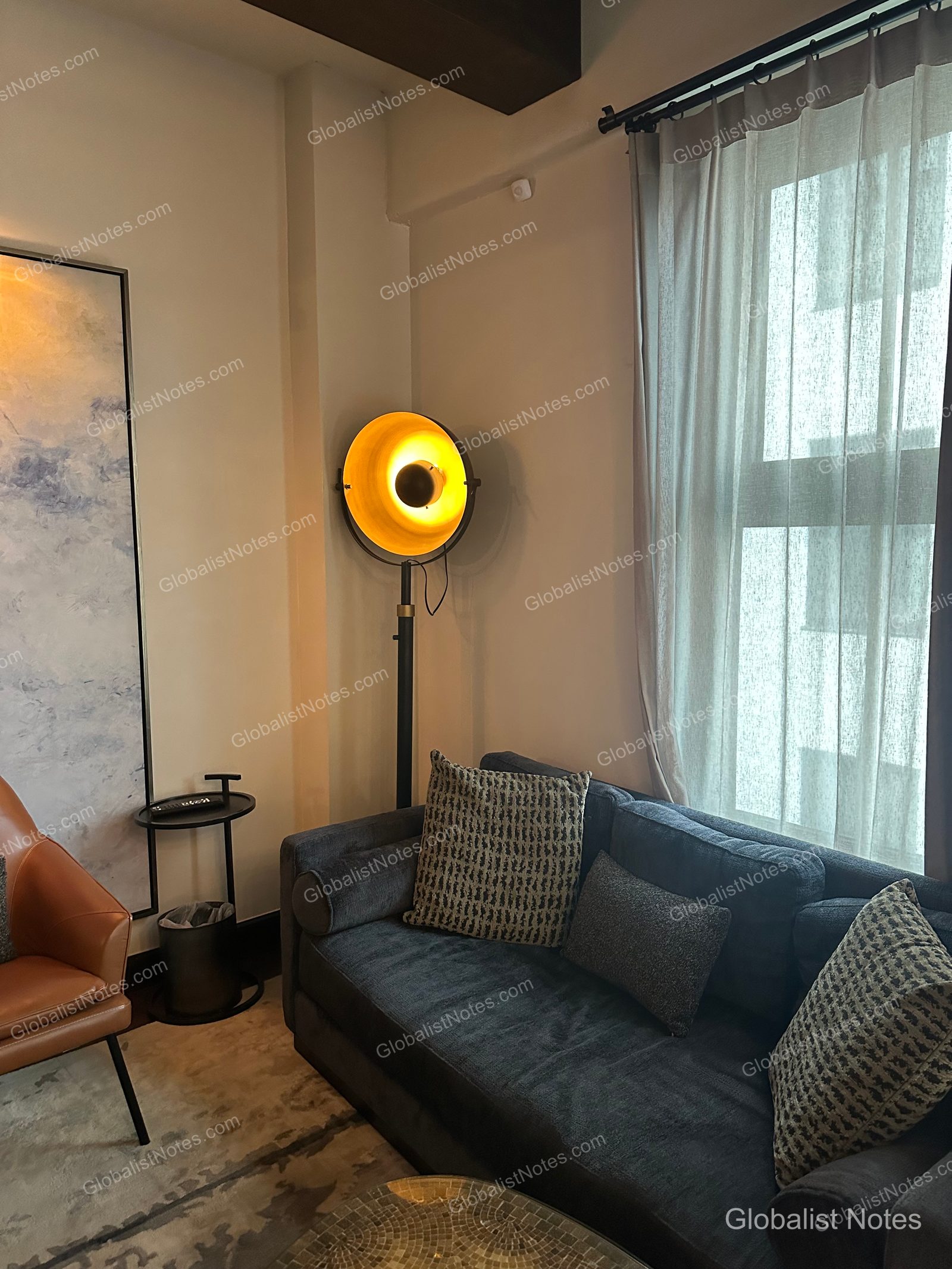 Living area of a Junior Suite at Hotel Figueroa in Los Angeles, featuring a dark gray sofa, a unique circular floor lamp, and abstract wall art. Sheer curtains cover the window, with a portion of a brown leather chair visible to the side.
