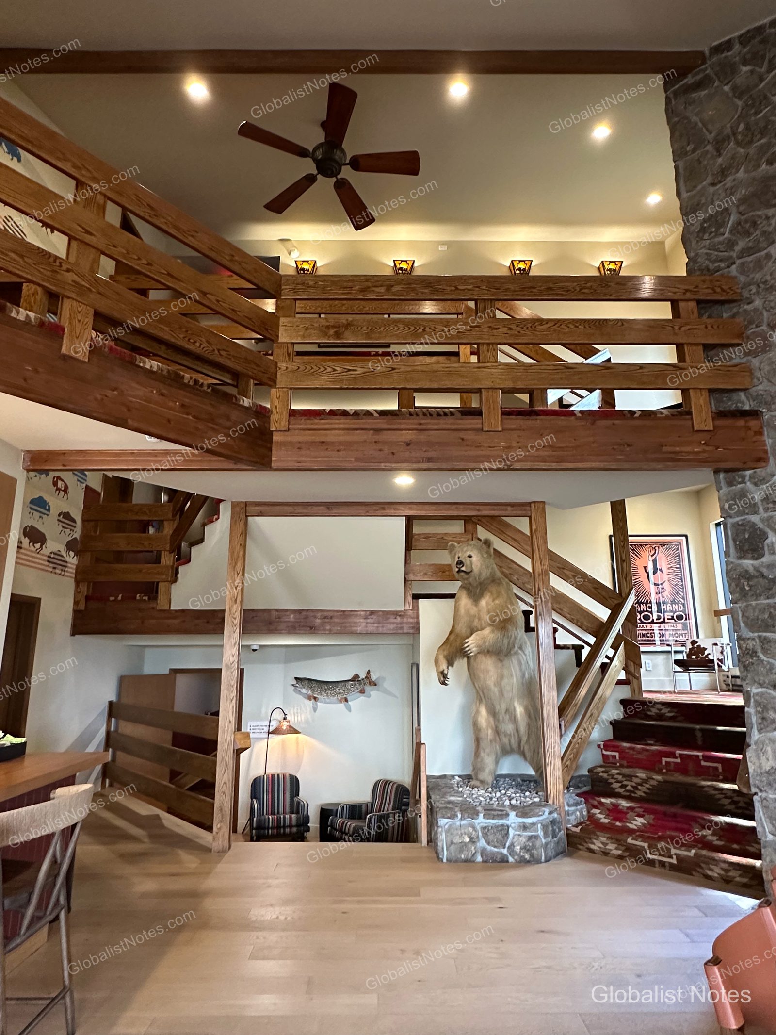 米国ボーズマン、Wandering Buffaloホテルのロビー。吹き抜けの2階建て構造で、中央にクマの剥製が置かれ、階段には柄入りのカーペットが敷かれています。壁面の魚の装飾やロデオのポスターが、山小屋のような雰囲気を演出しています。