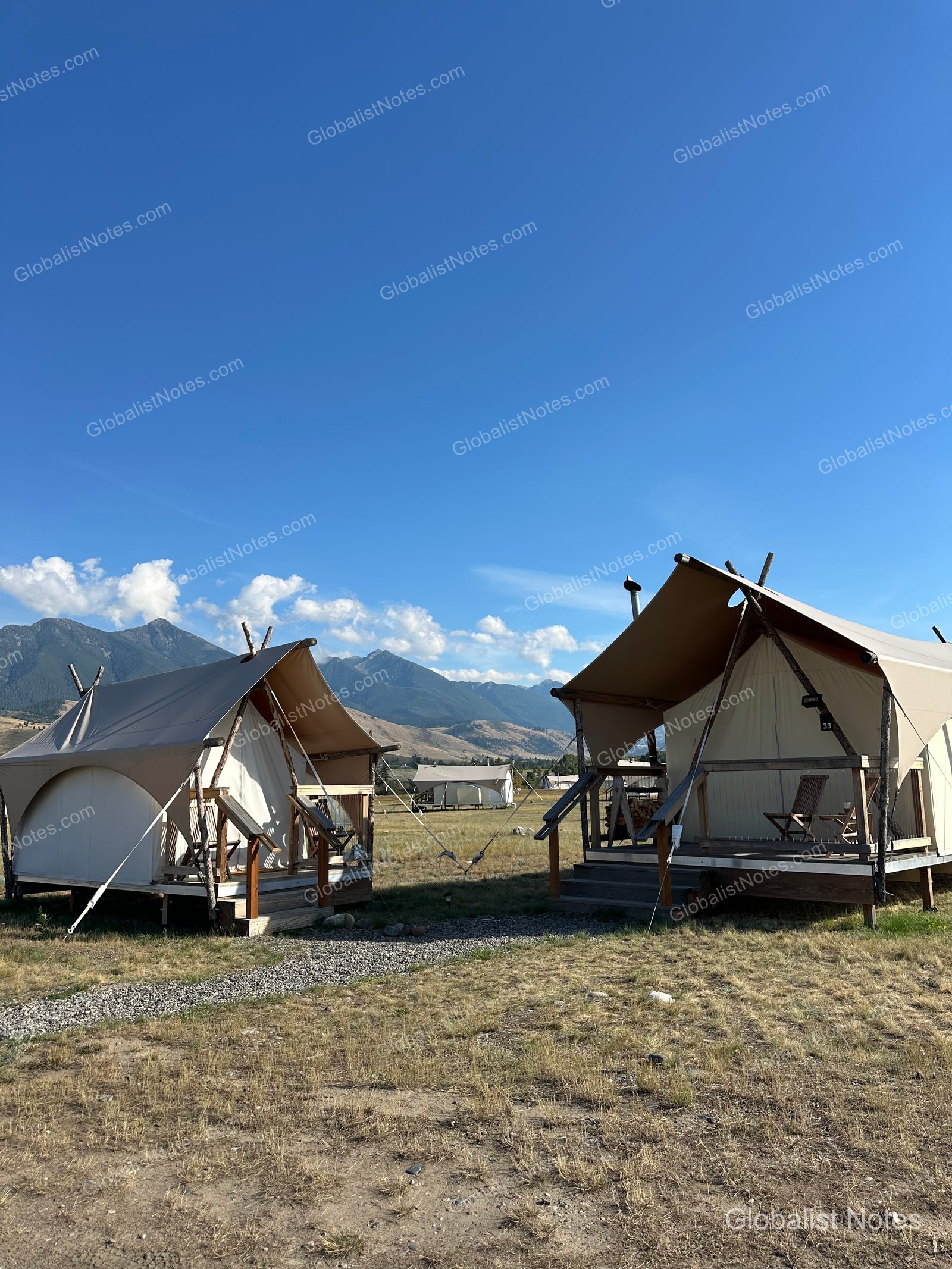 Under Canvas North Yellowstone - Paradise Valleyのテント群。ウッドデッキを備えたキャンバステントが草原に並び、背後には山脈と青空が広がっています。
