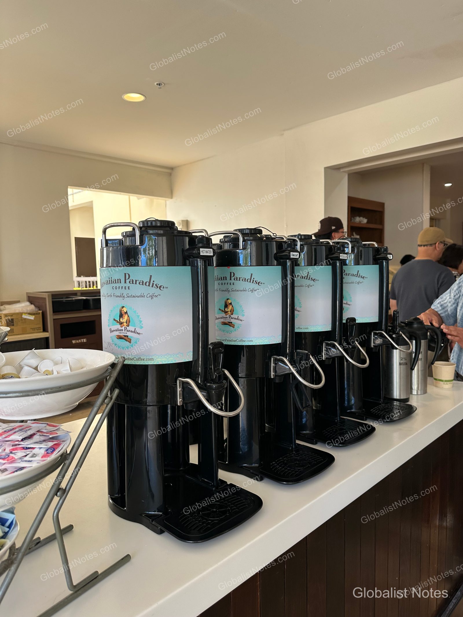 Four black Hawaiian Paradise coffee dispensers lined up on a white counter in the breakfast area at Hyatt Place Waikiki Beach, with bowls of sugar and cream nearby.
