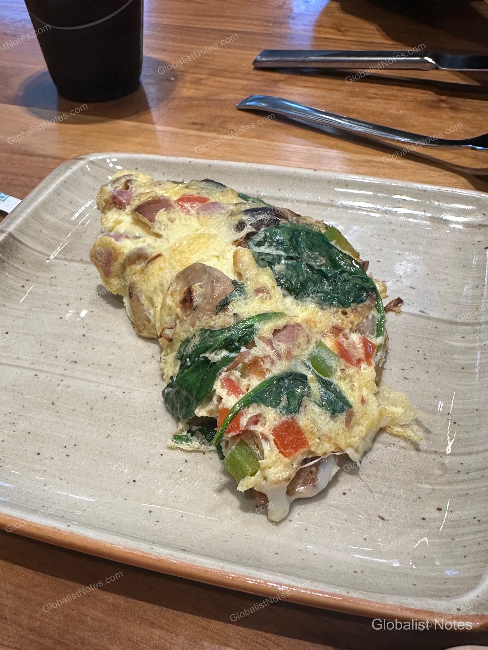An omelet with spinach, tomatoes, and bacon served on a light beige square plate, with a fork, knife, and black beverage cup placed on a wooden table.