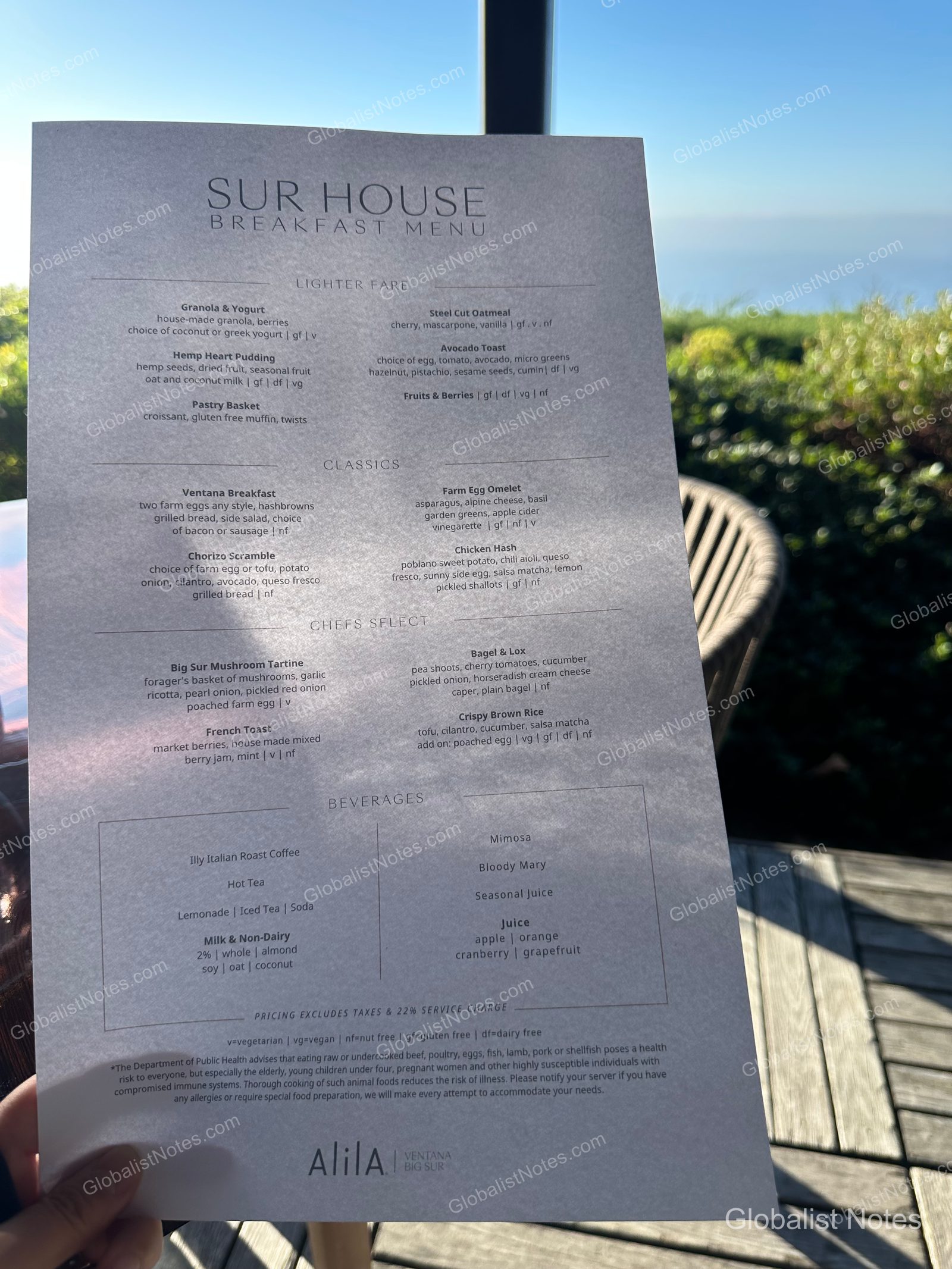The Sur House breakfast menu at Alila Ventana Big Sur placed on an outdoor table. The menu lists items such as granola, avocado toast, and the Ventana breakfast, with the blue ocean and trees of Big Sur blurred in the background.