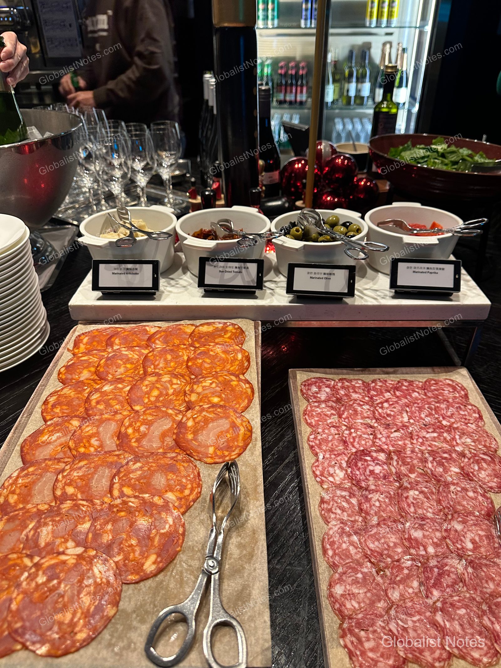 A buffet table during the evening cocktail hour at the Grand Hyatt Jeju lounge. Large platters of sliced ham are displayed alongside small bowls of olives, pickled vegetables, and cheese, with a refrigerator containing wine and beverages visible in the background.