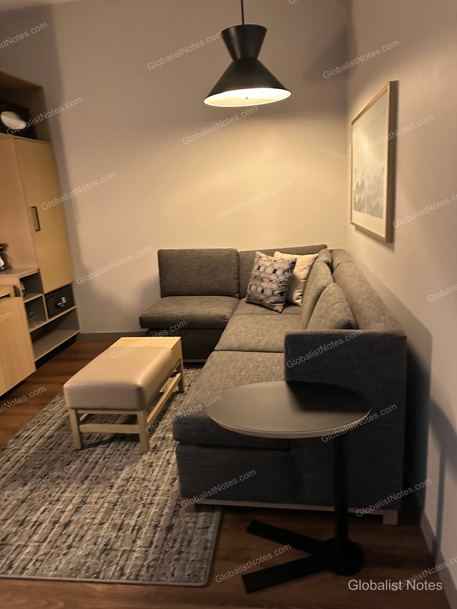 hyatt place Bozeman Yellowstone Airportのジュニアスイートのリビングエリア。グレーのL字型ソファ、ベージュのオットマン、小さな円形サイドテーブルが置かれ、壁にはアート、天井にはペンダントライトが配されている。