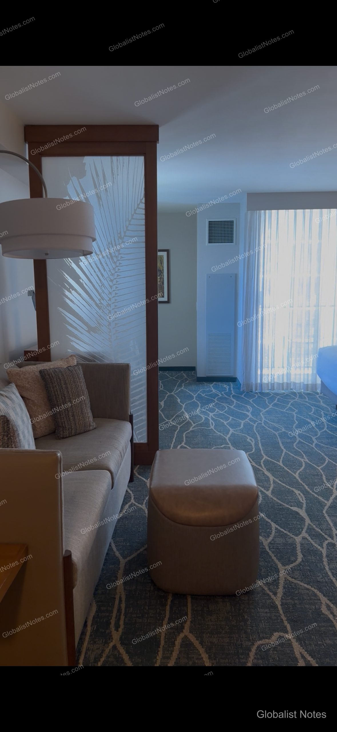 Interior of a guest room at Hyatt Place Waikiki Beach, showing a living area with a sofa and ottoman, a translucent partition with palm leaf patterns, and carpeted flooring leading toward the window.