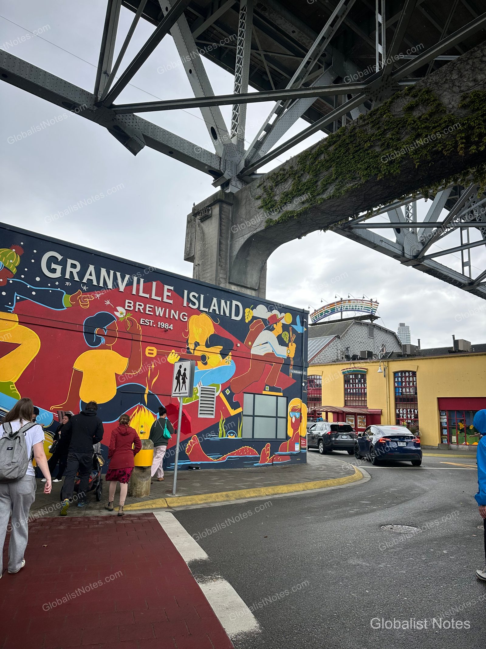 Colorful murals on the Granville Island Brewing building in Vancouver, with people walking beneath the massive steel bridge structure overhead, nearby traffic, and a yellow building in the background.